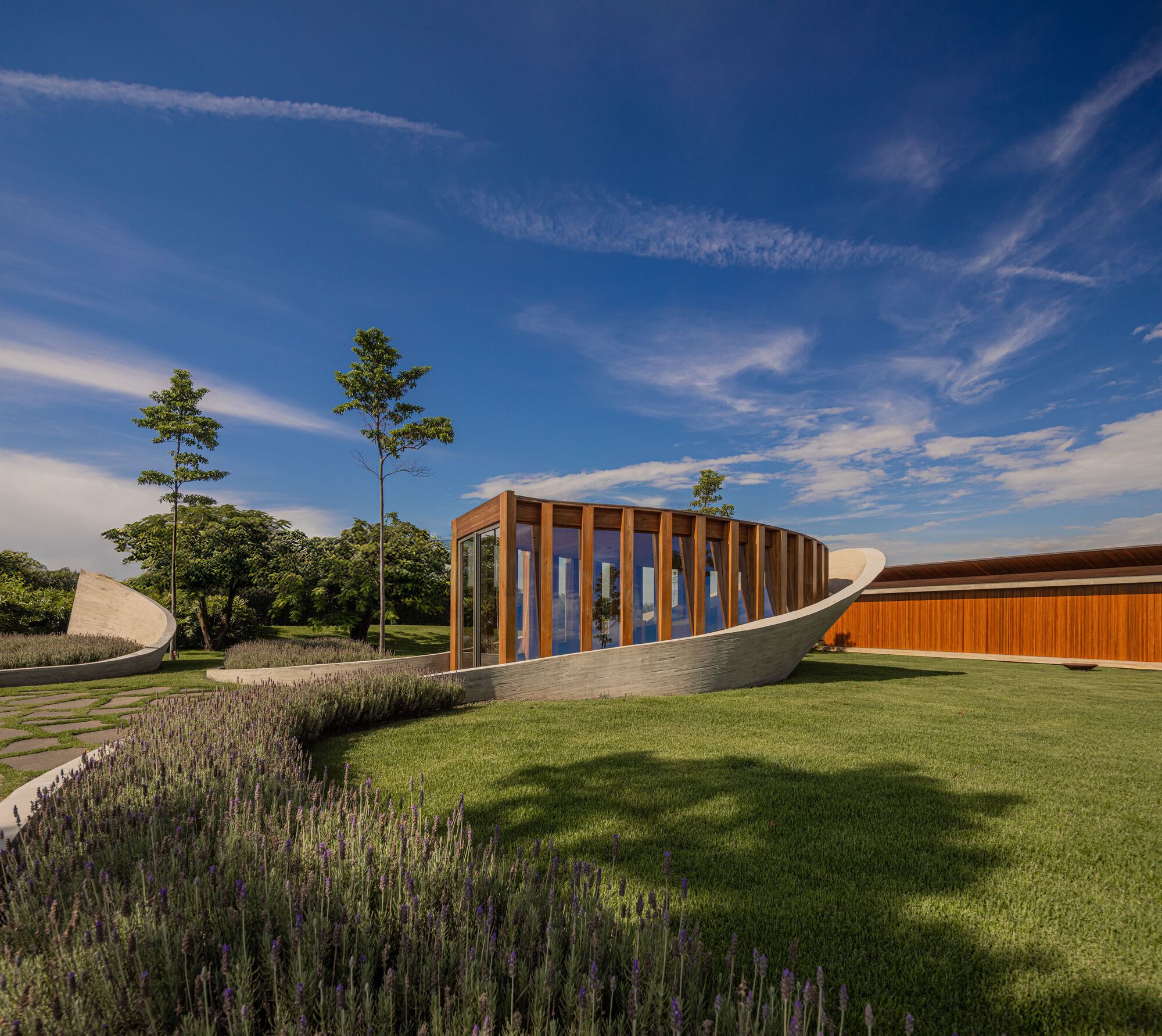 Curving white concrete path with planted lavender beds leading to timber-clad glazed pavilion under blue sky