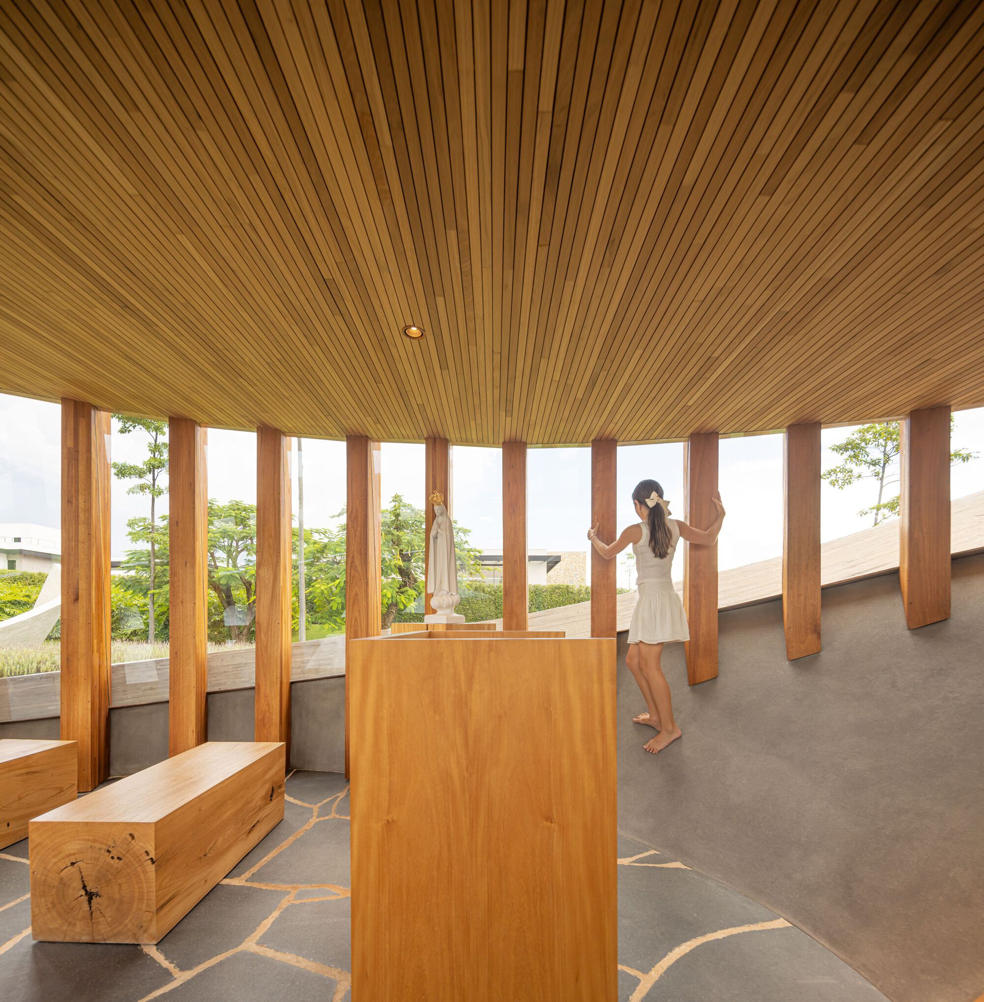 Radial timber ceiling and column-divided windows overlooking planted terrace while a woman stands barefoot inside