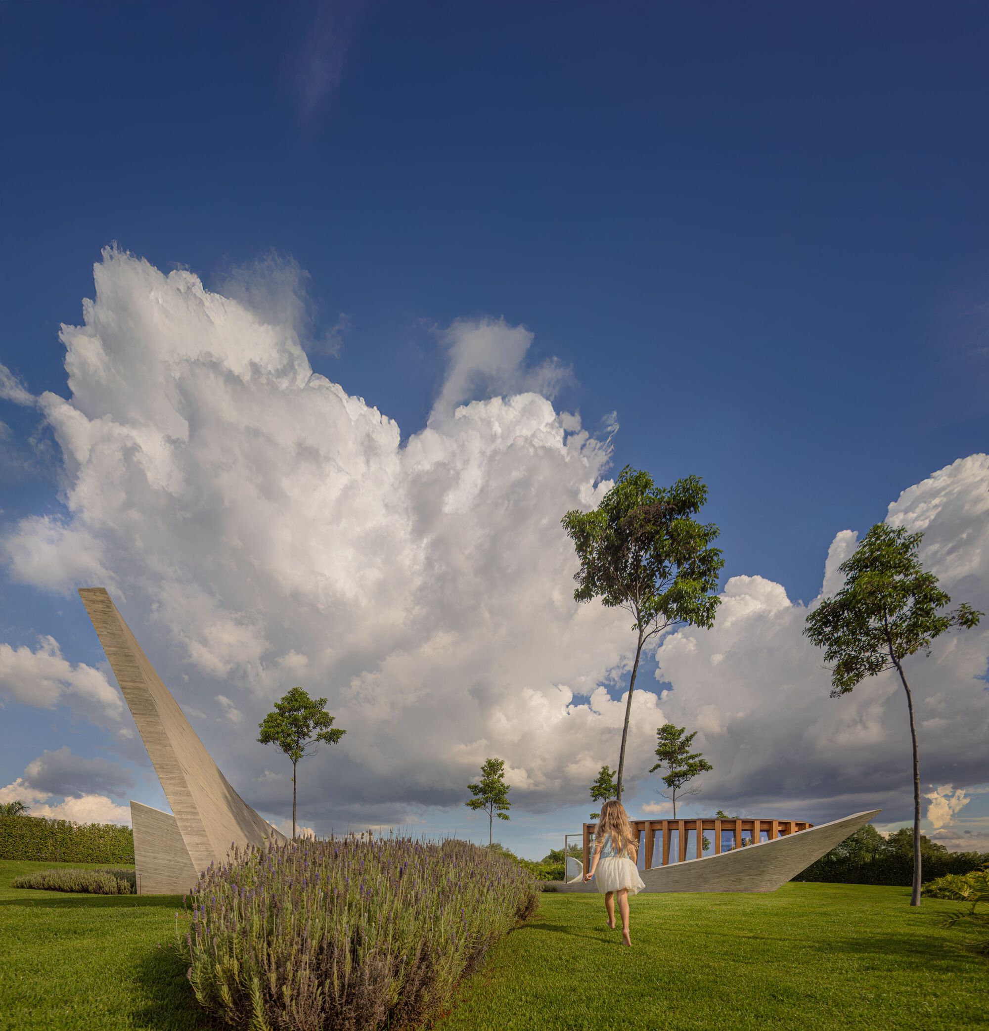 Curved concrete walls and timber colonnade framing the lawn under towering cumulonimbus clouds
