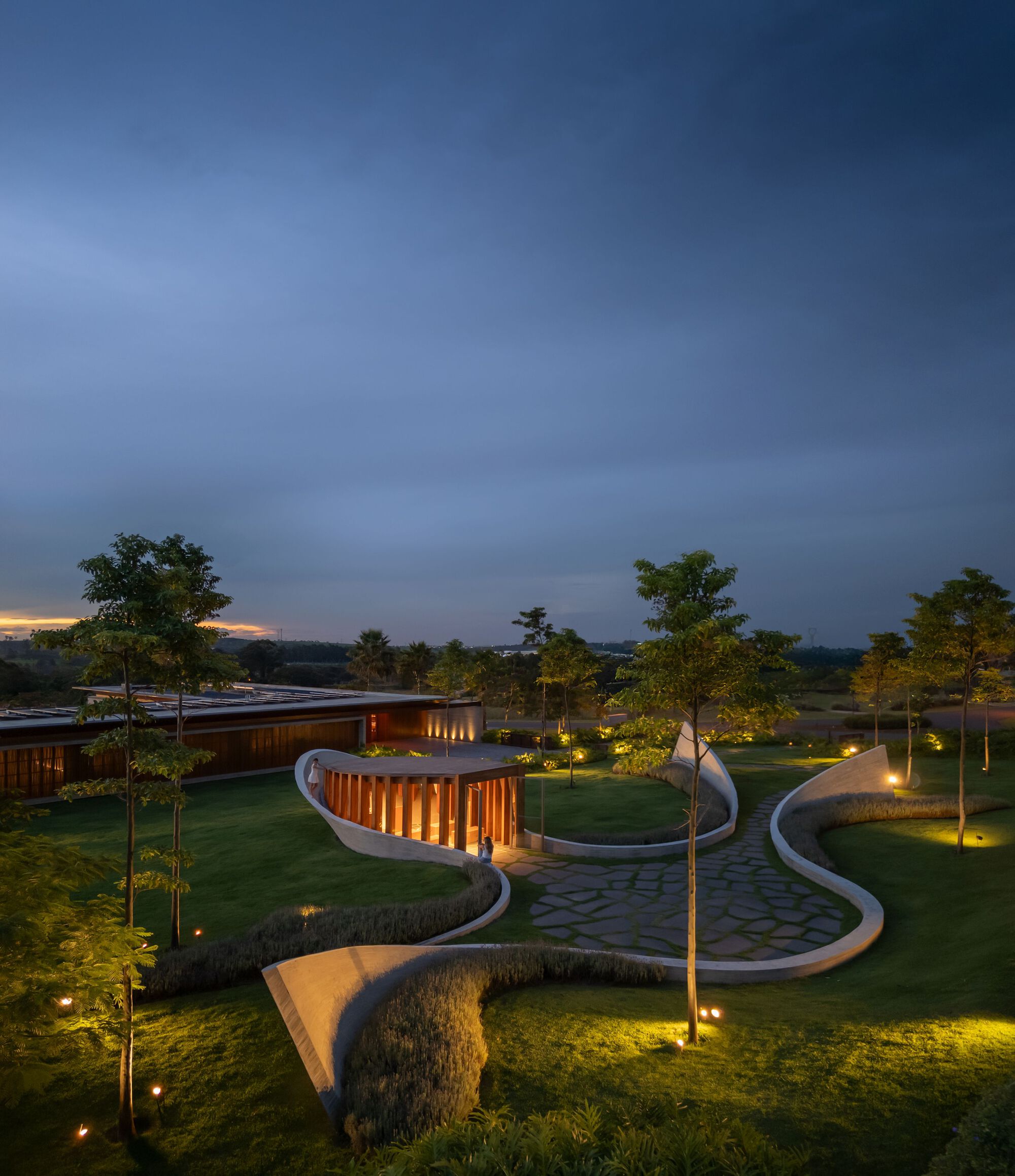 Curving landscape walls with vertical timber screens and uplighting among the lawn at dusk