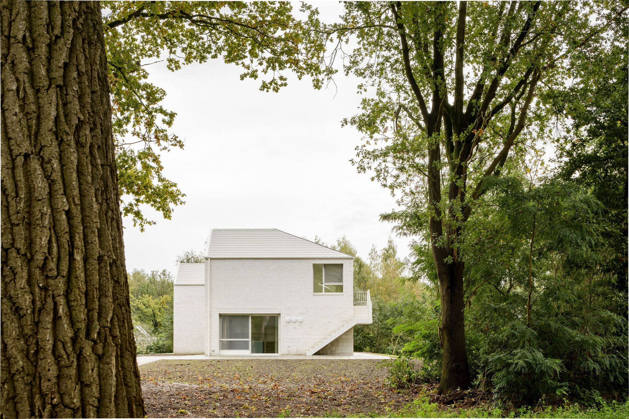Rear elevation of white brick volumes with gabled roofs seen through mature deciduous trees
