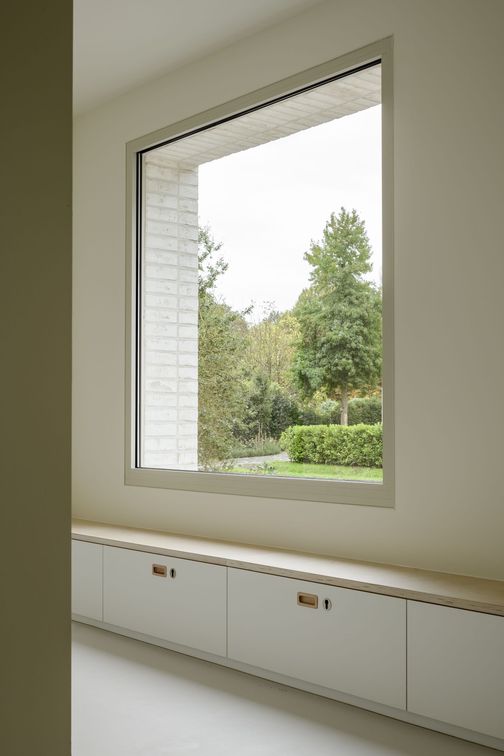 Interior window seat with built-in drawers overlooking green garden through deep brick reveal