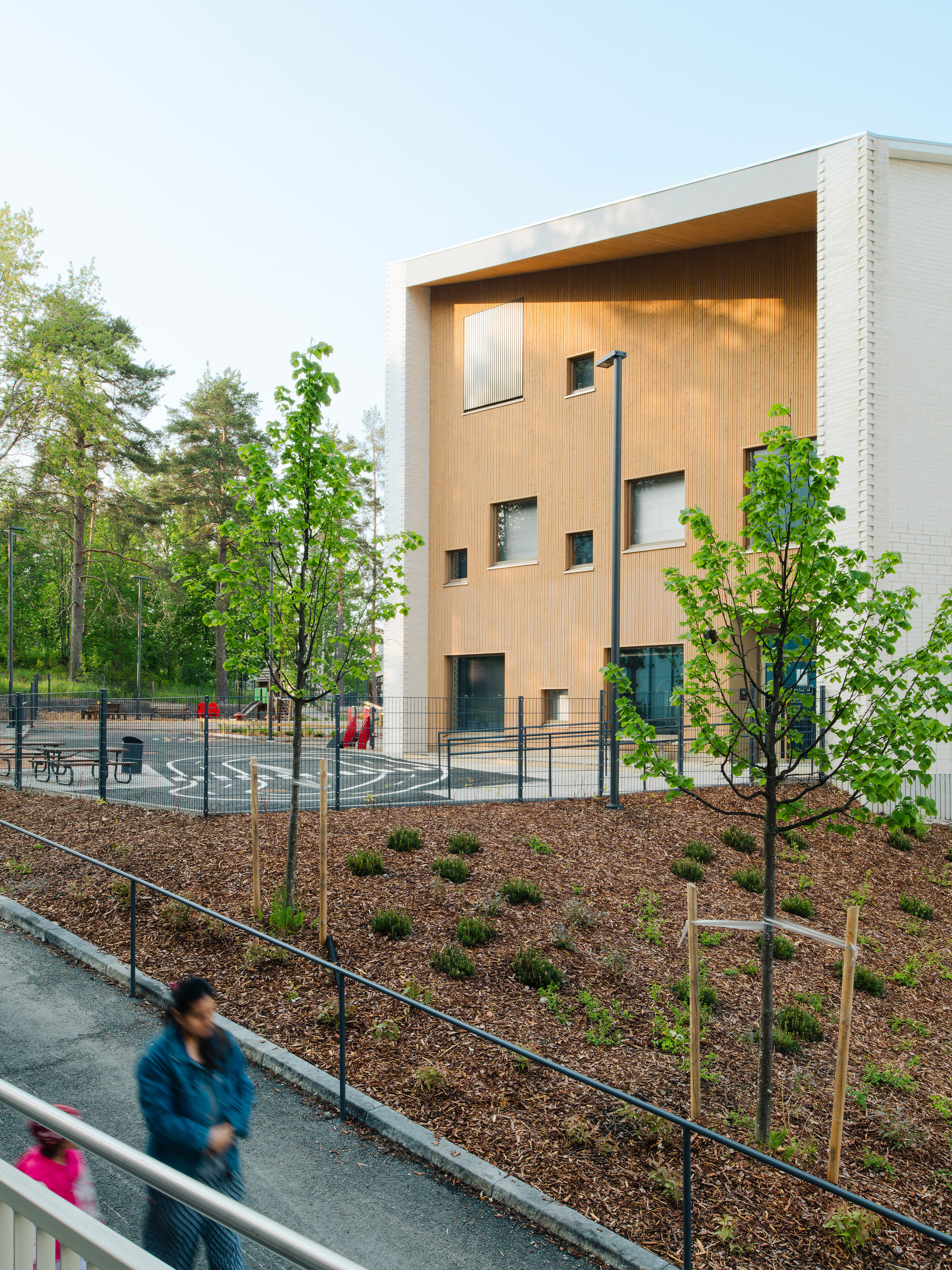 Timber-clad facade with scattered windows above a sloped planted bed and a figure passing below