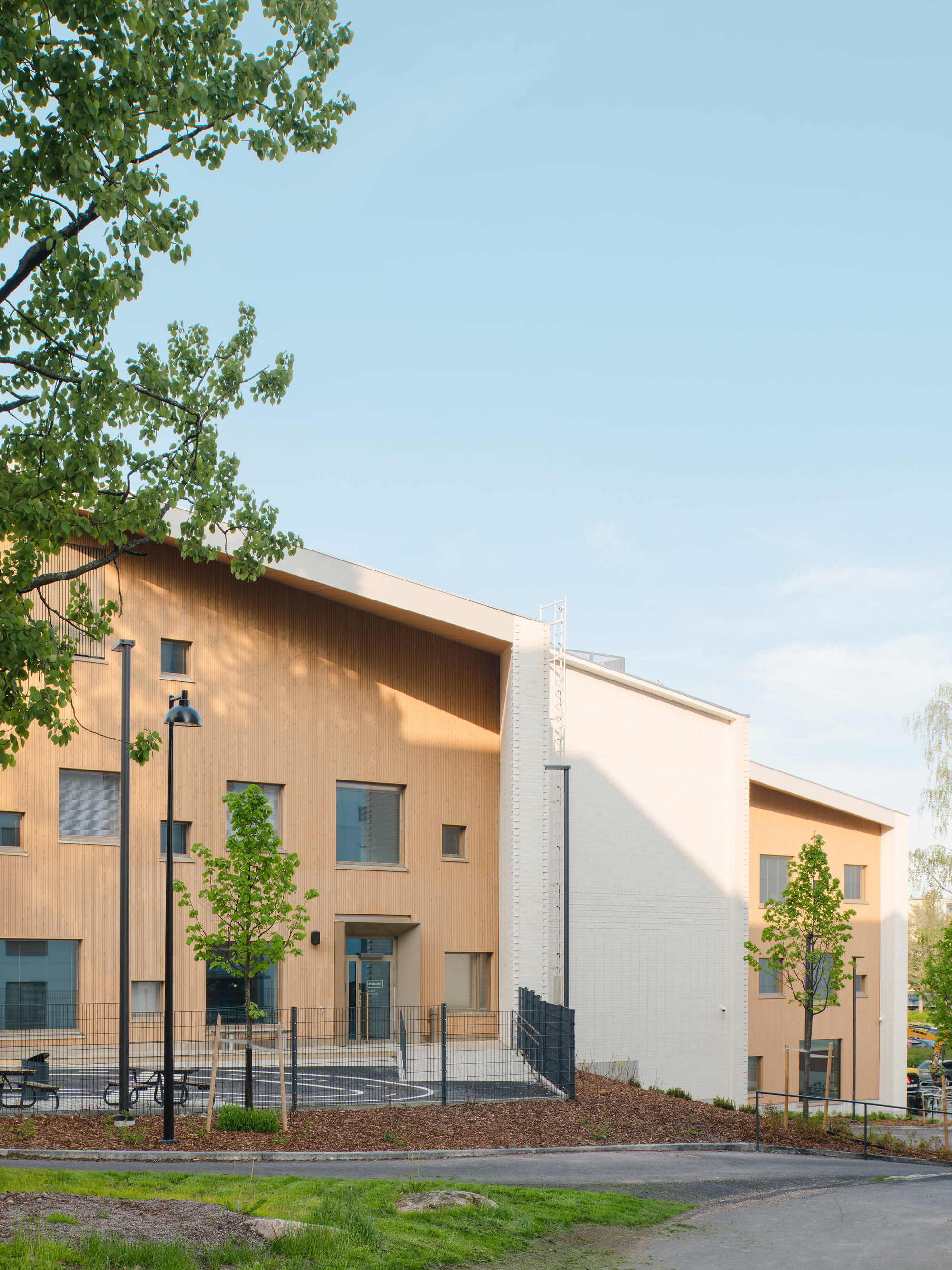 Street view of the timber-clad entrance facade with young trees in late afternoon light