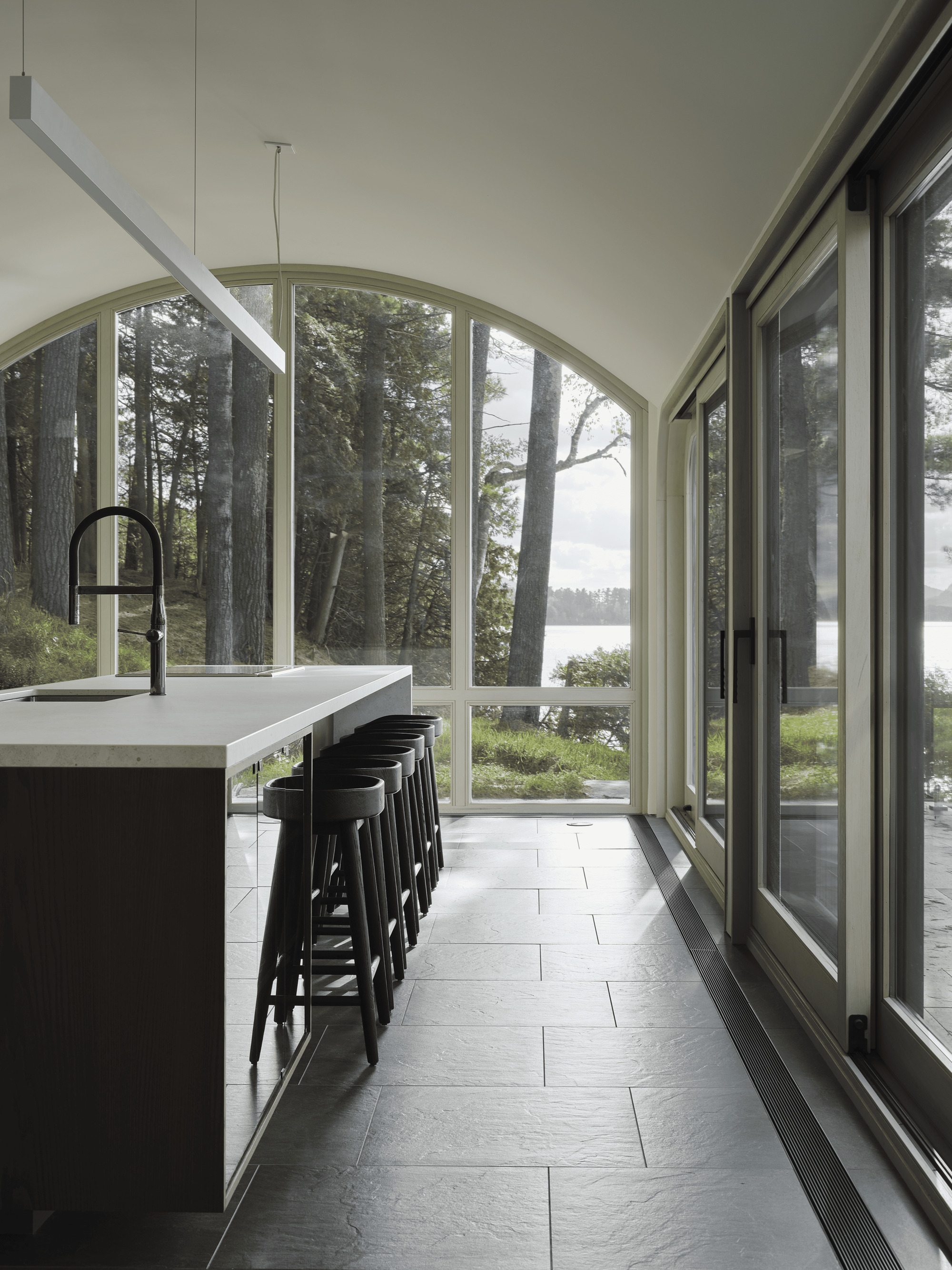 Kitchen island with black stools beneath arched windows framing pine trees and water beyond