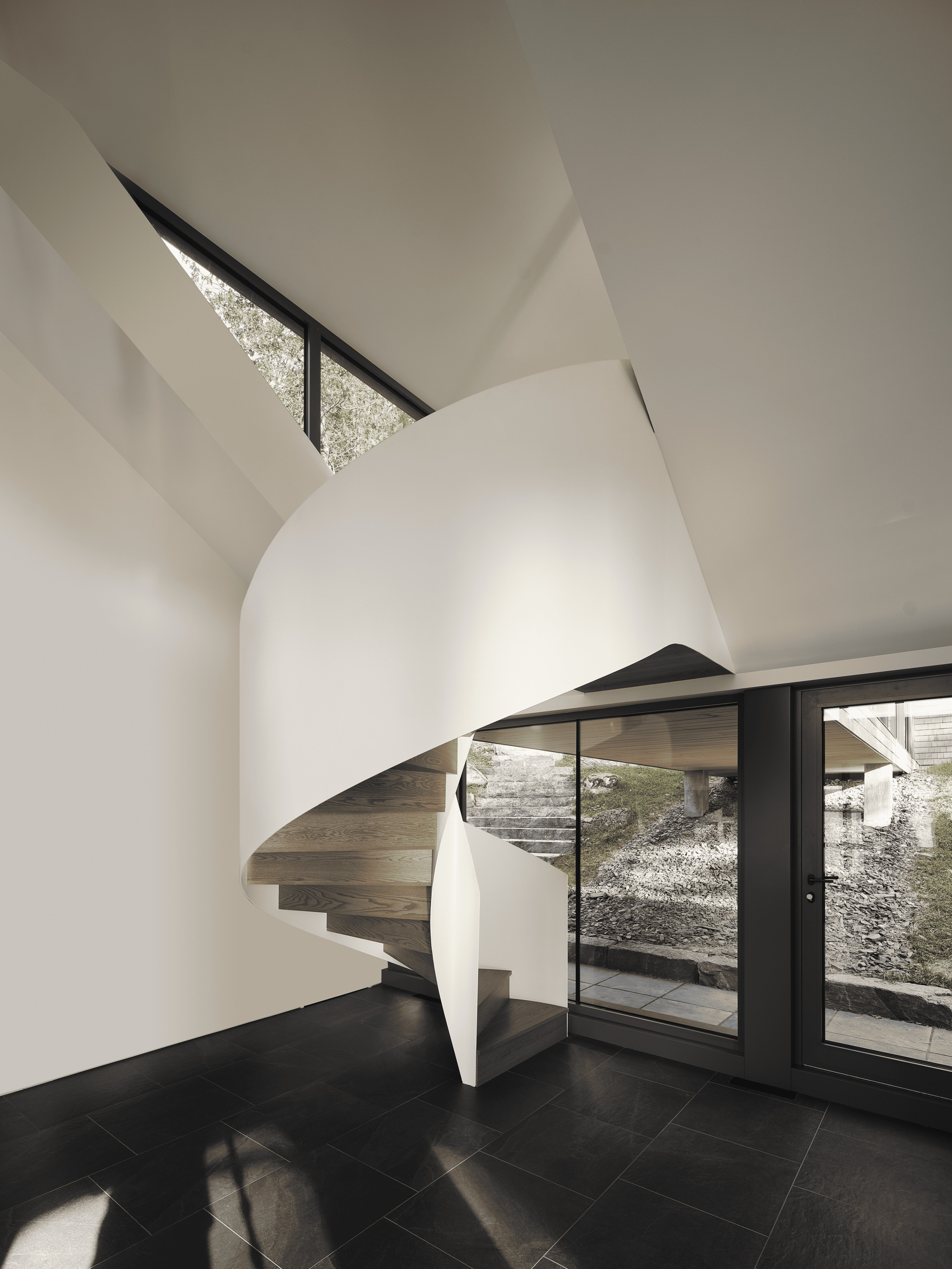 Curved sculptural staircase wrapped in white walls beside dark-framed glass doors opening to courtyard