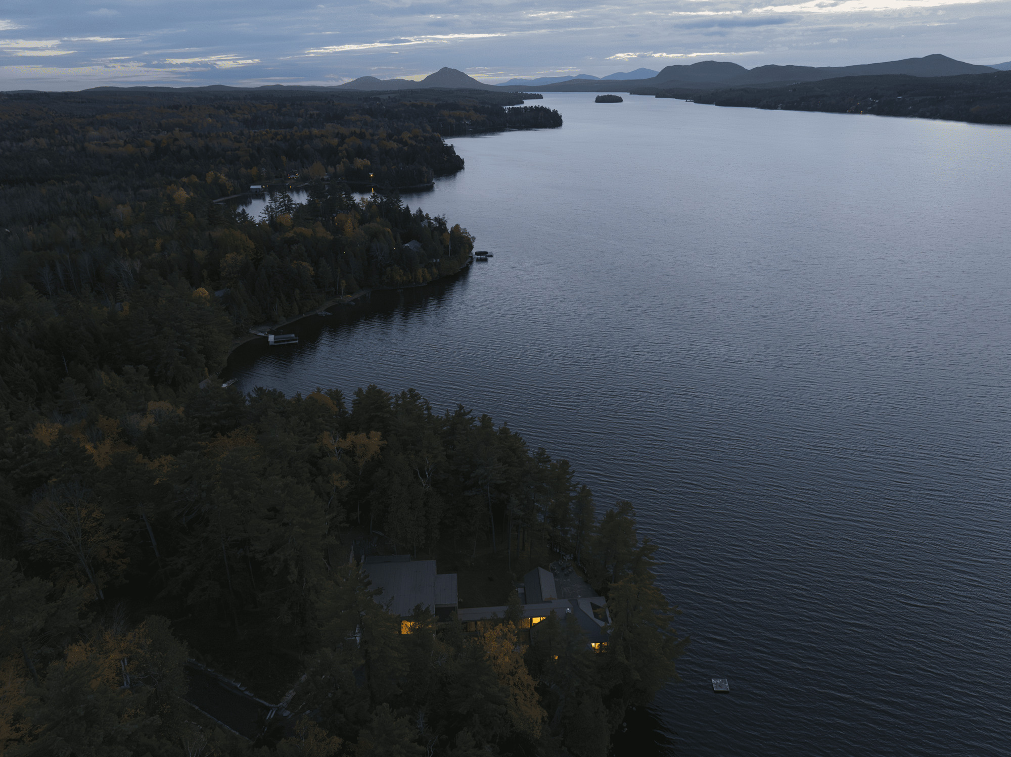 Aerial view of a lakeside residence nestled among dense forest along a curving shoreline at dusk