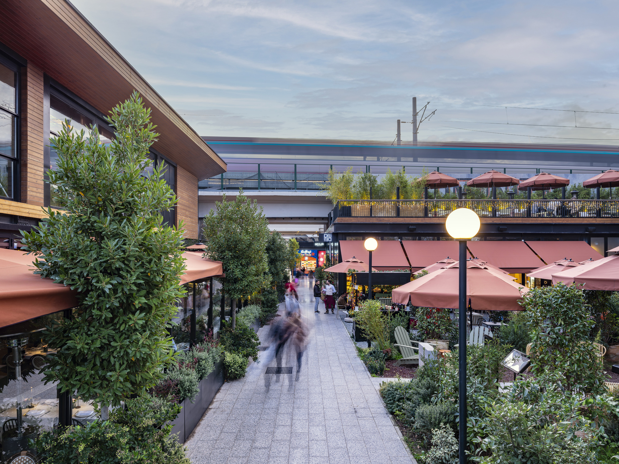 Pedestrian promenade lined with planters and cafe umbrellas beneath the elevated rail line at dusk