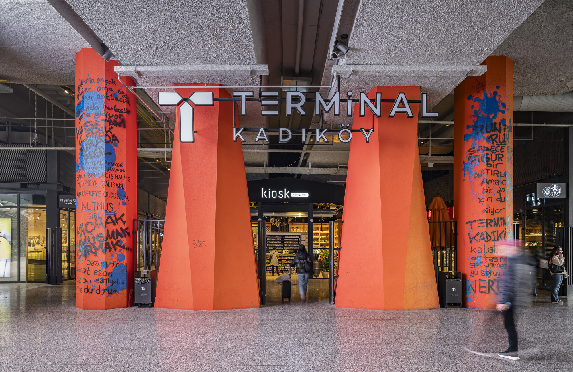 Four tapered orange columns supporting the concrete canopy with pedestrians passing through the concourse