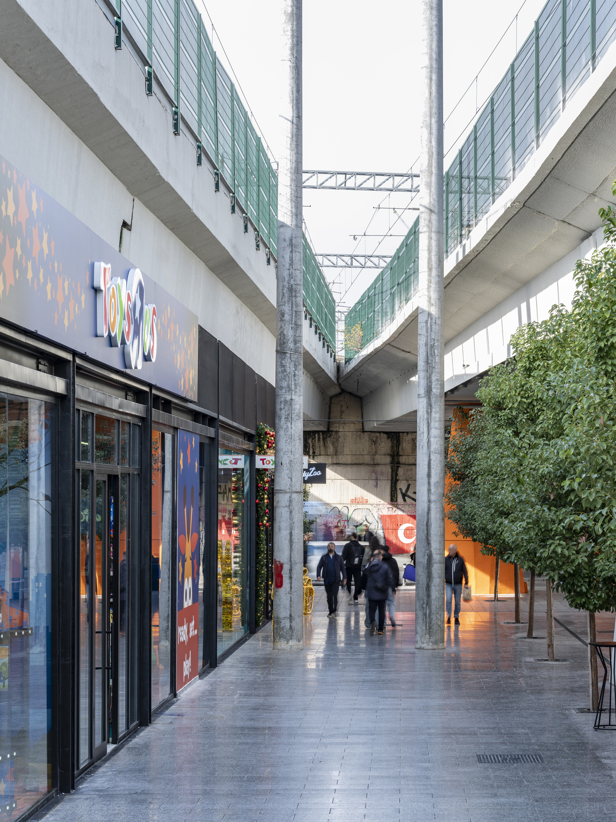 Glazed corridor beneath the concrete viaduct with retail storefronts and pedestrians on wet pavement