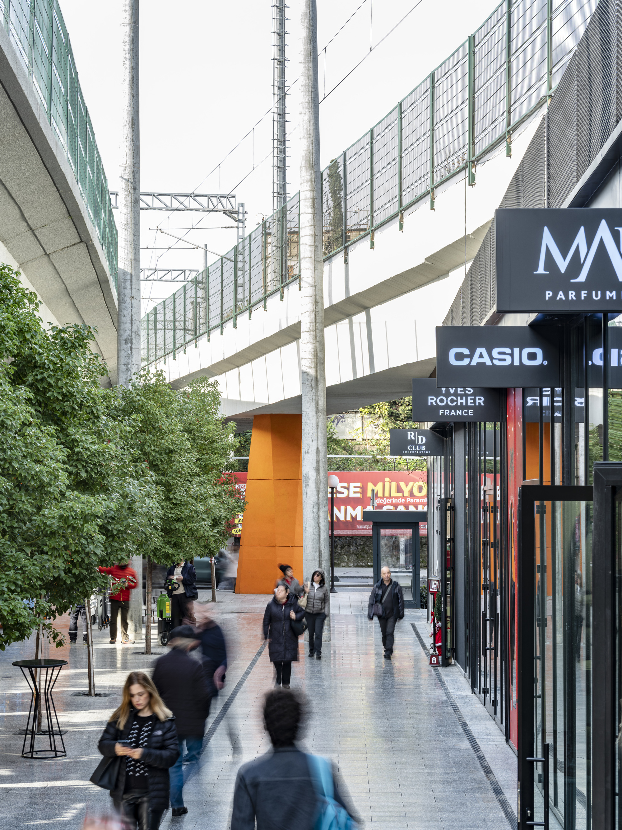Shopping passage with polycarbonate screen walls and planted trees between retail facades at dusk