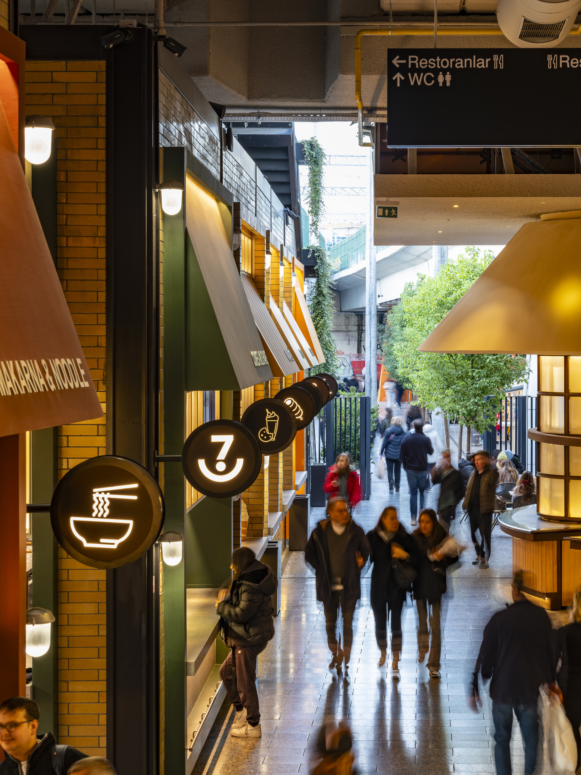 Food hall corridor with angled awnings and circular signage as visitors walk toward daylit courtyard