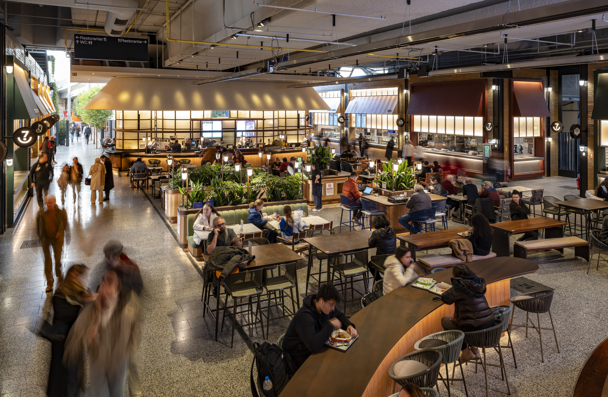 Central food hall seating area with communal tables beneath illuminated kiosk canopies and exposed ceiling systems