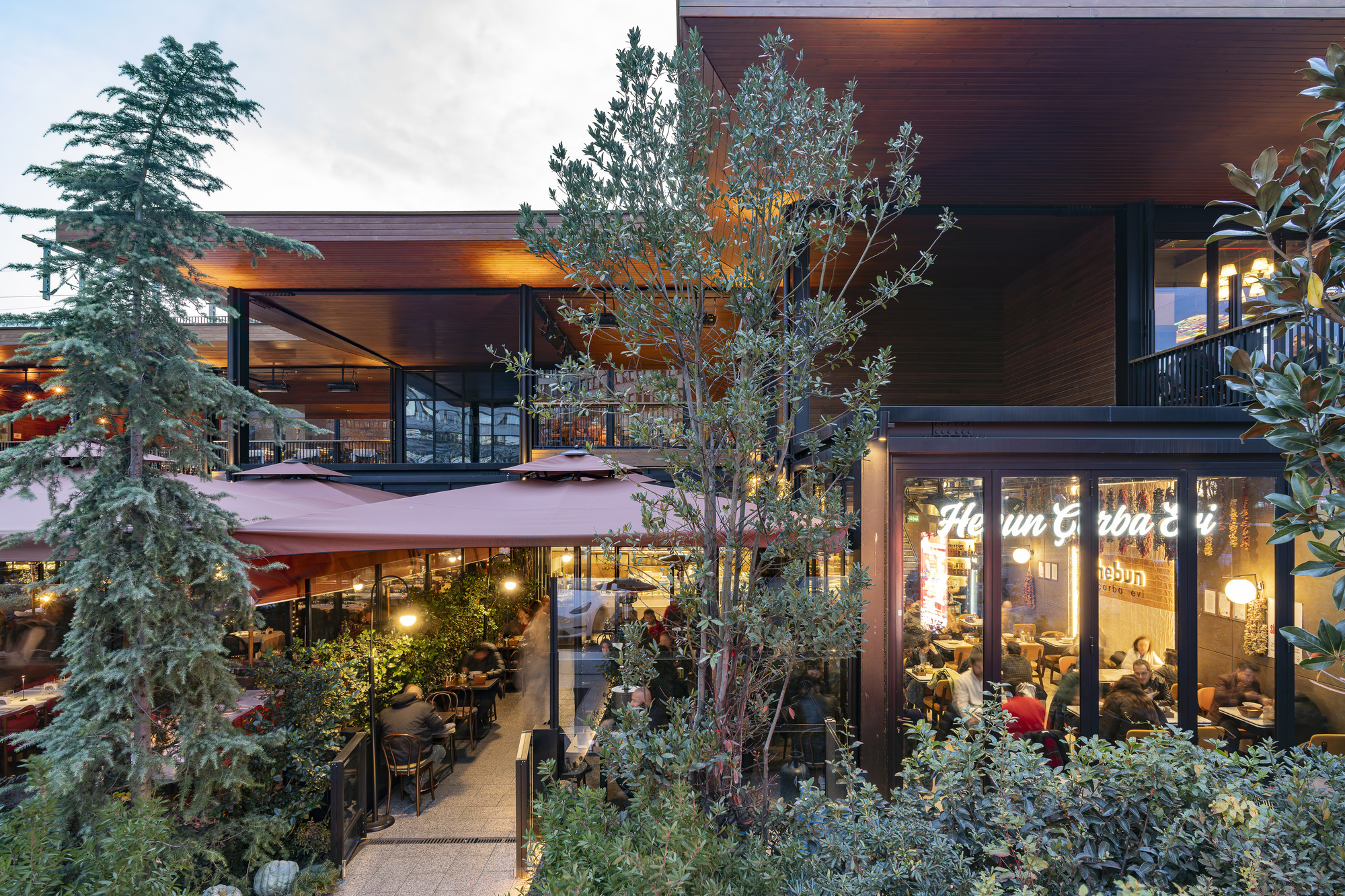 Two-level timber-clad structure with planted terraces and glass-enclosed dining areas at dusk