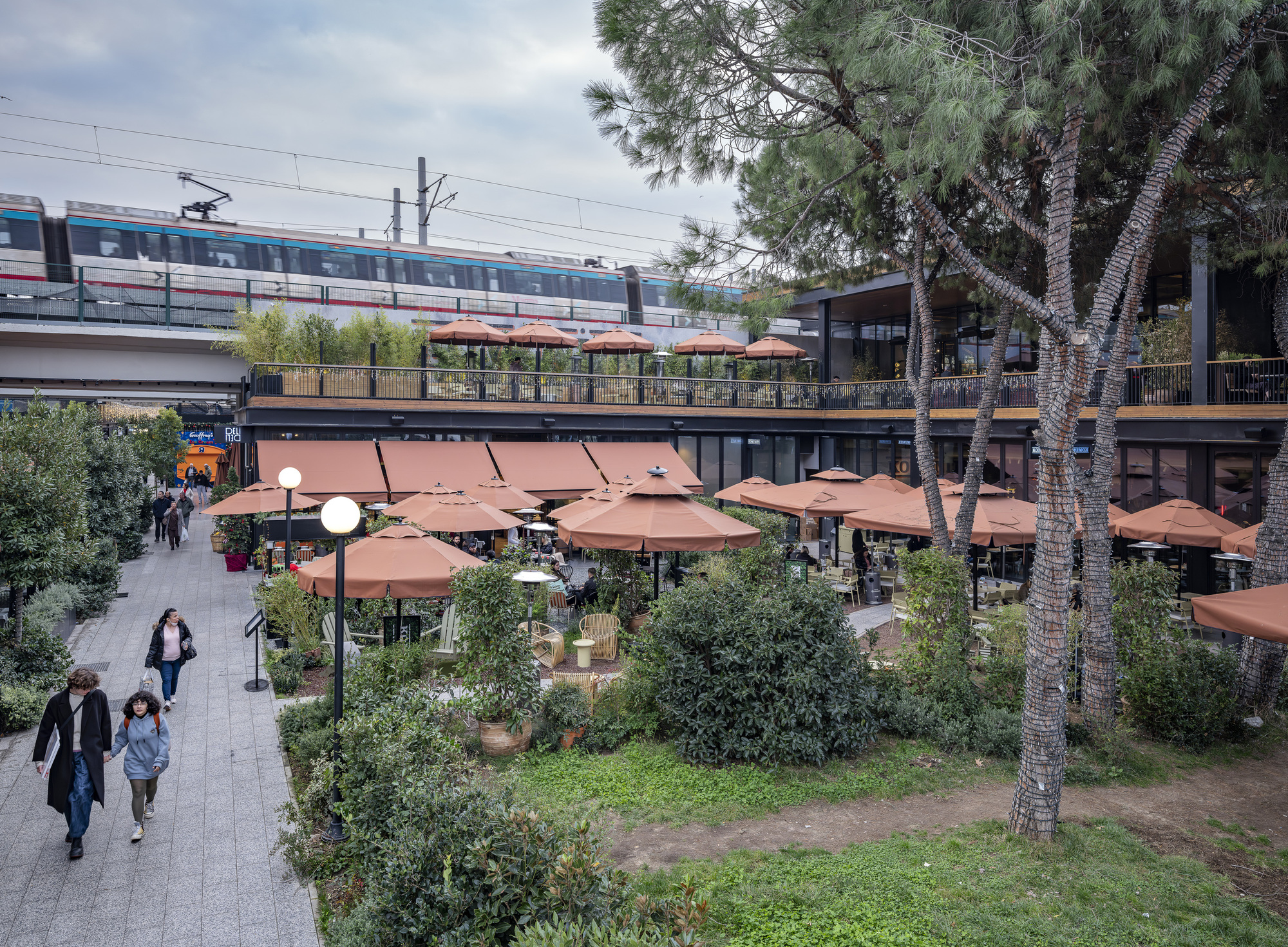 Open courtyard with terracotta umbrellas and pedestrians walking beneath an elevated train passing overhead