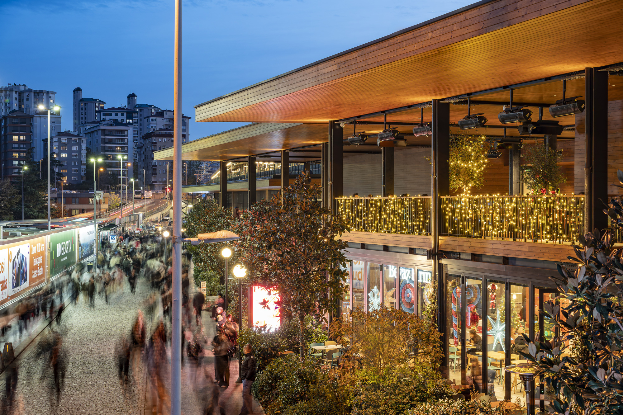 Timber-lined canopy extending over a two-story glass facade with string lights and evening crowd below