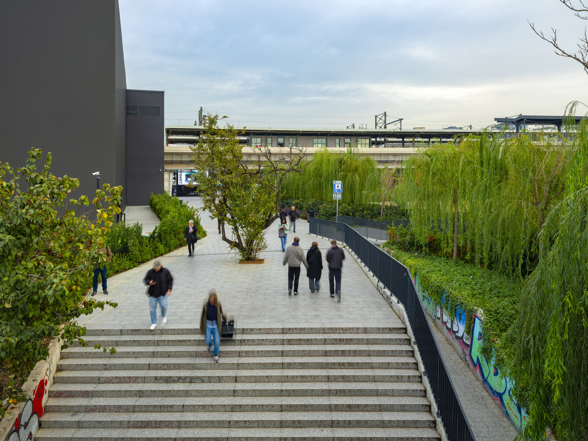 Stone staircase descending to a planted pedestrian plaza with people walking under cloudy skies