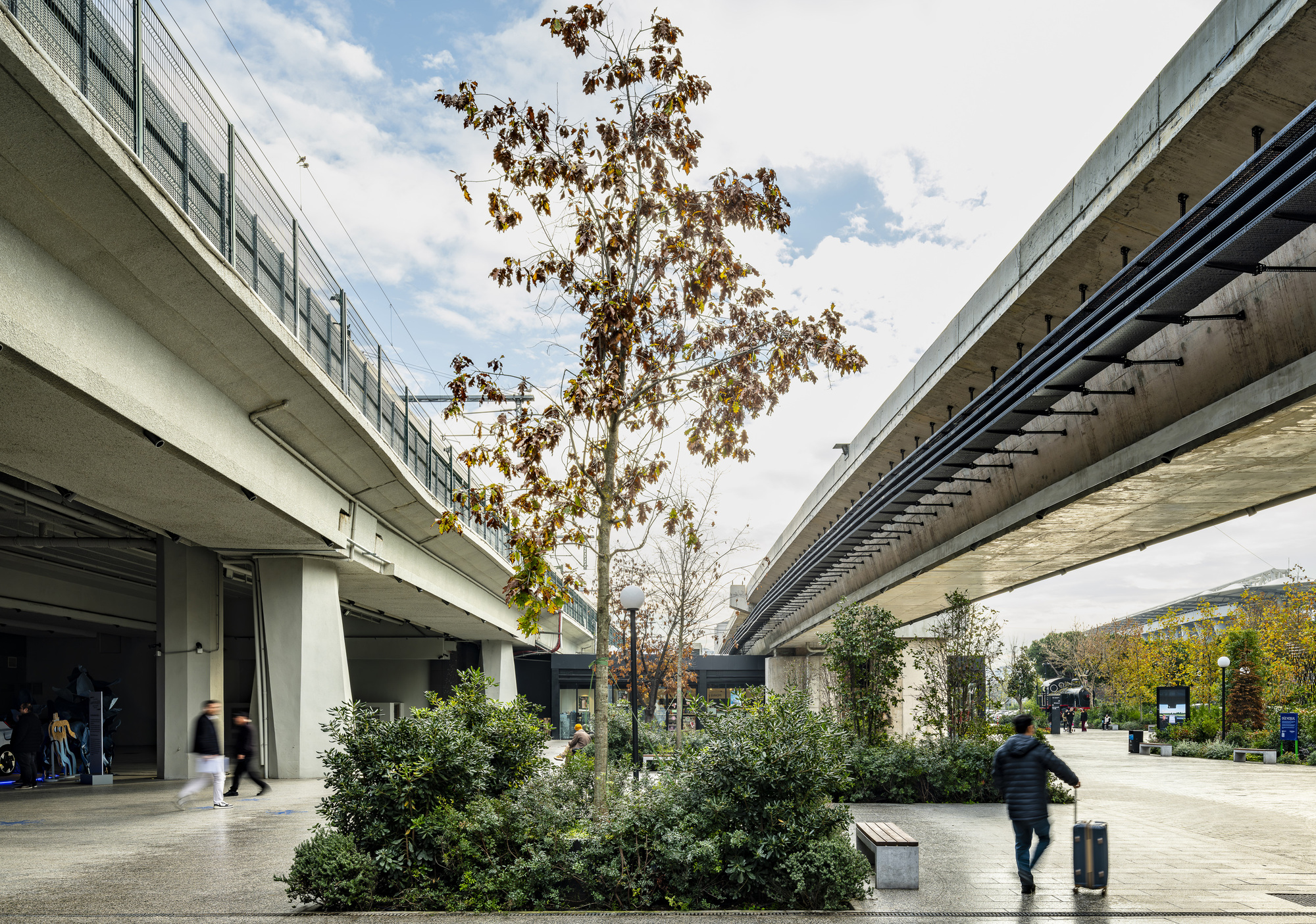 Concrete transit viaducts flanking a planted courtyard with a person walking past shrubs in afternoon light
