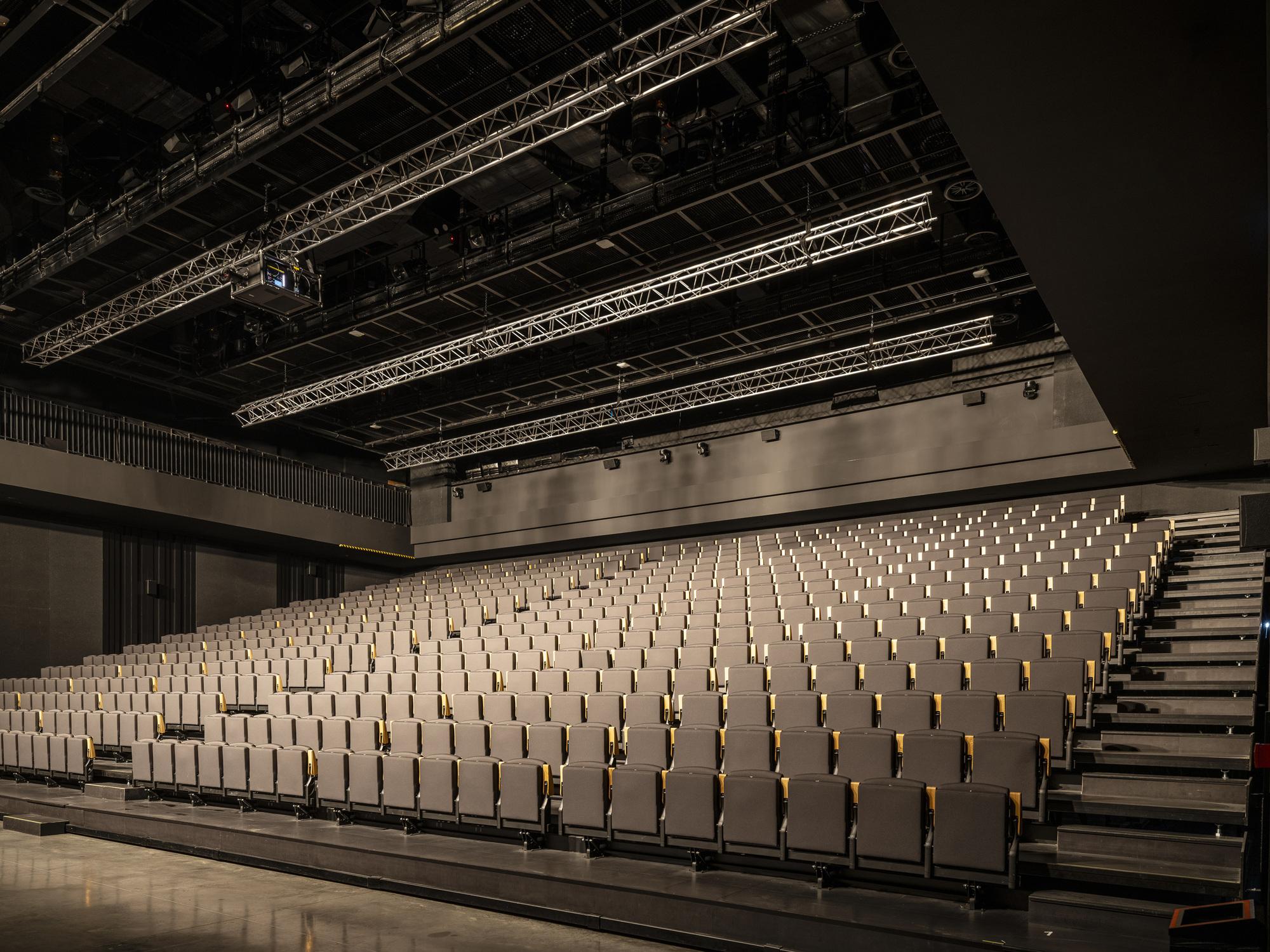 Auditorium with raked timber seating benches, exposed metal truss ceiling, and theatrical lighting rig overhead