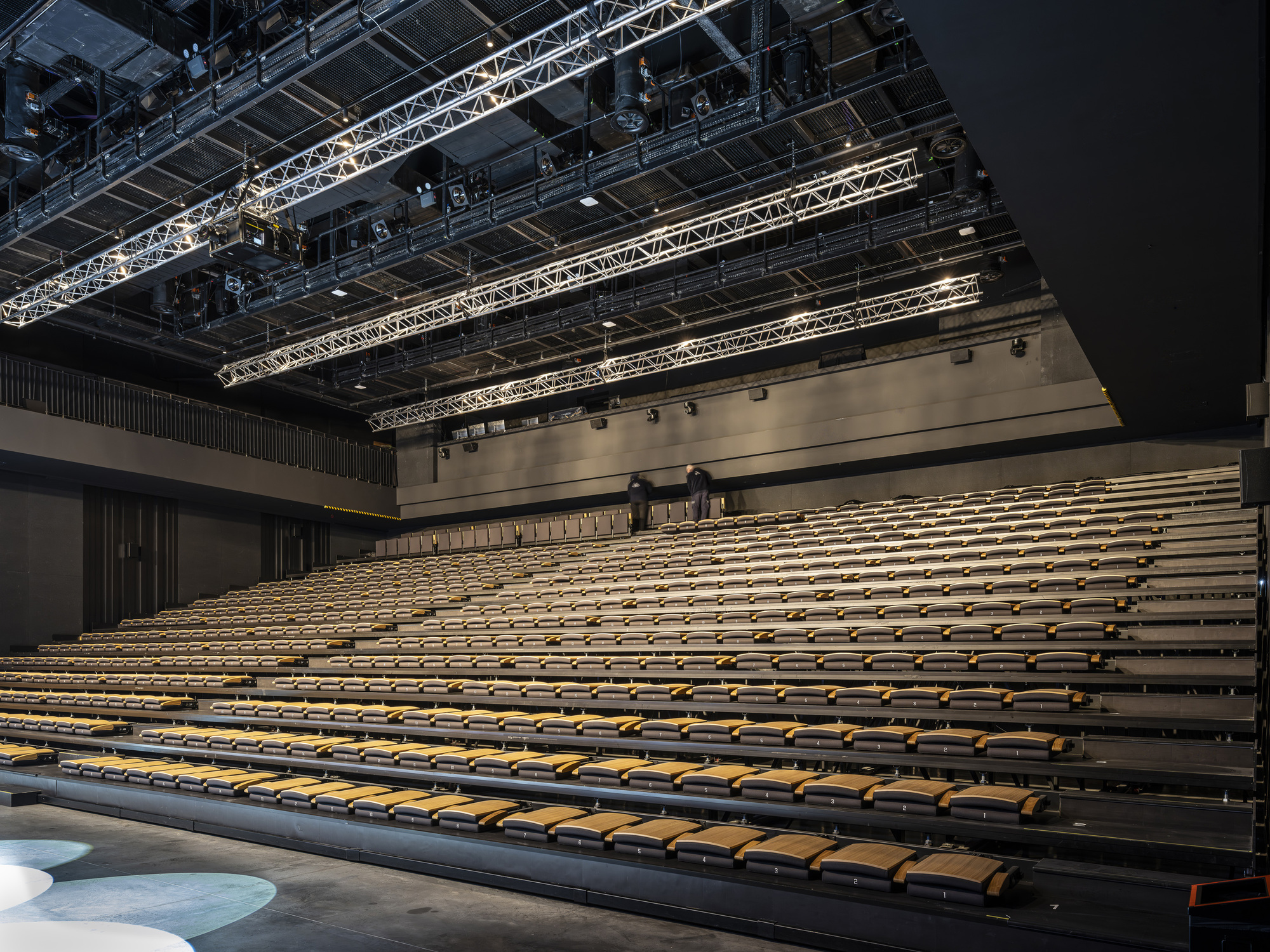 Auditorium seating with two figures standing at the upper tier of wooden benches under theatrical lighting
