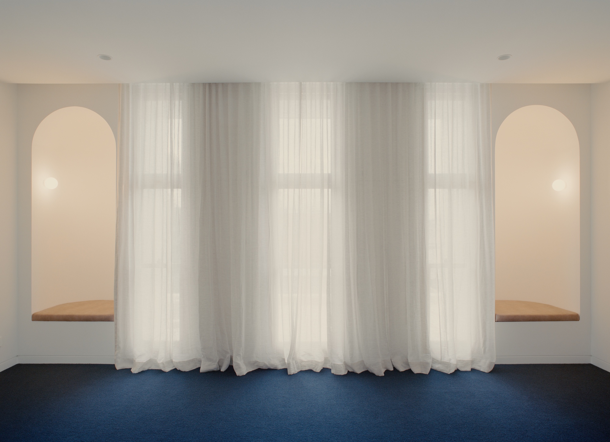 Symmetrical wall with twin arched niches and timber window seats behind floor-length sheer curtains over blue carpet