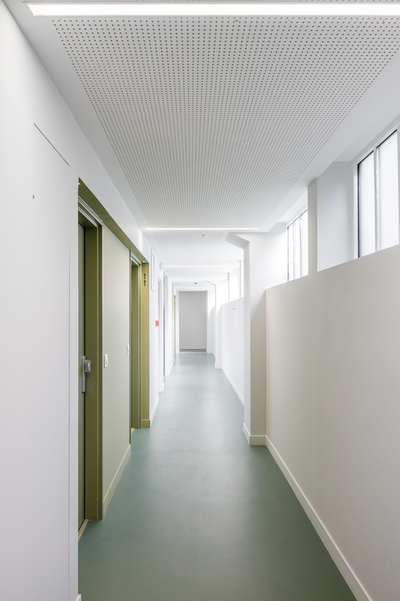 Corridor with perforated acoustic ceiling panels and green rubber flooring leading to olive-green unit doors