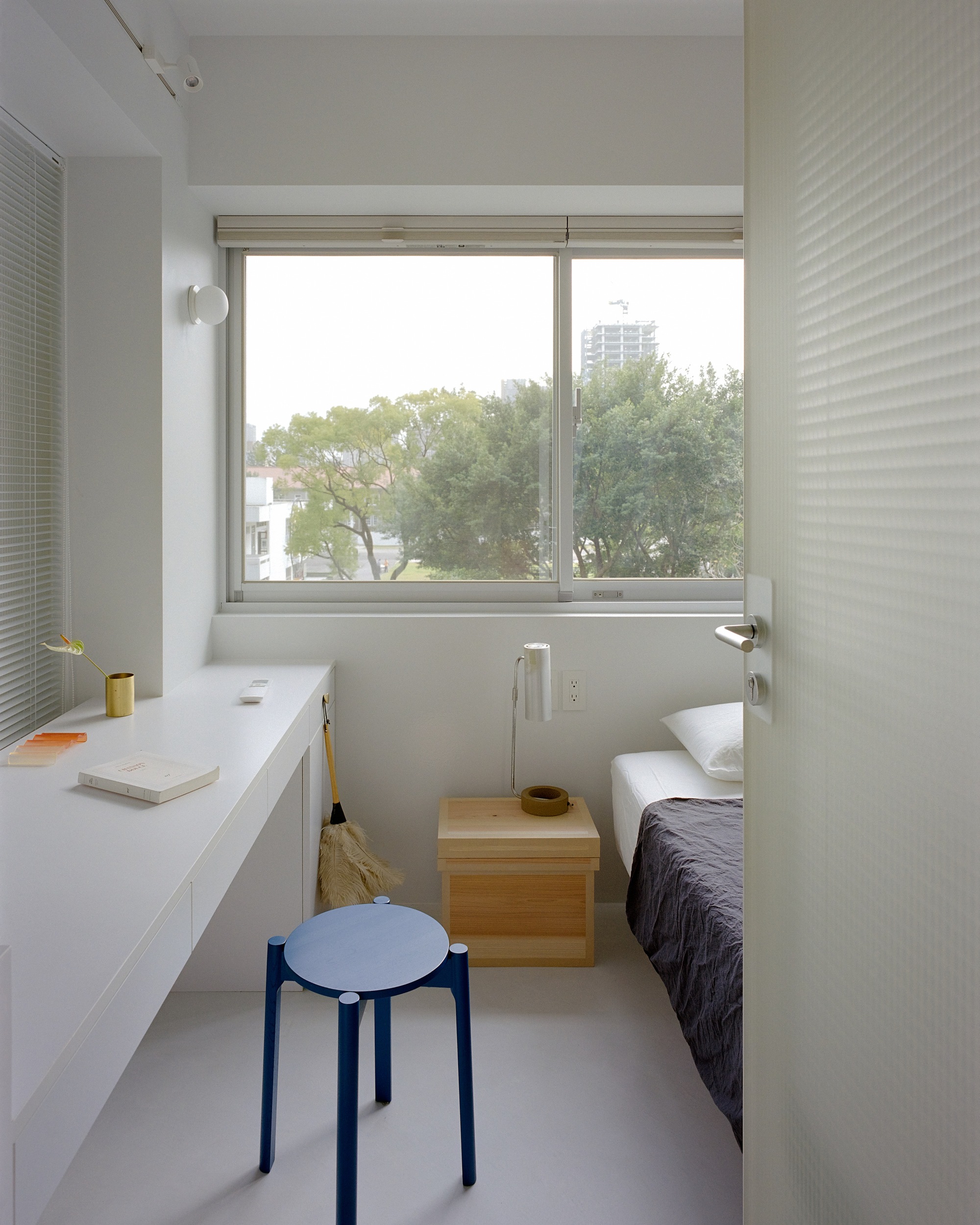 Compact bedroom with white desk, blue stool and window framing green trees outside