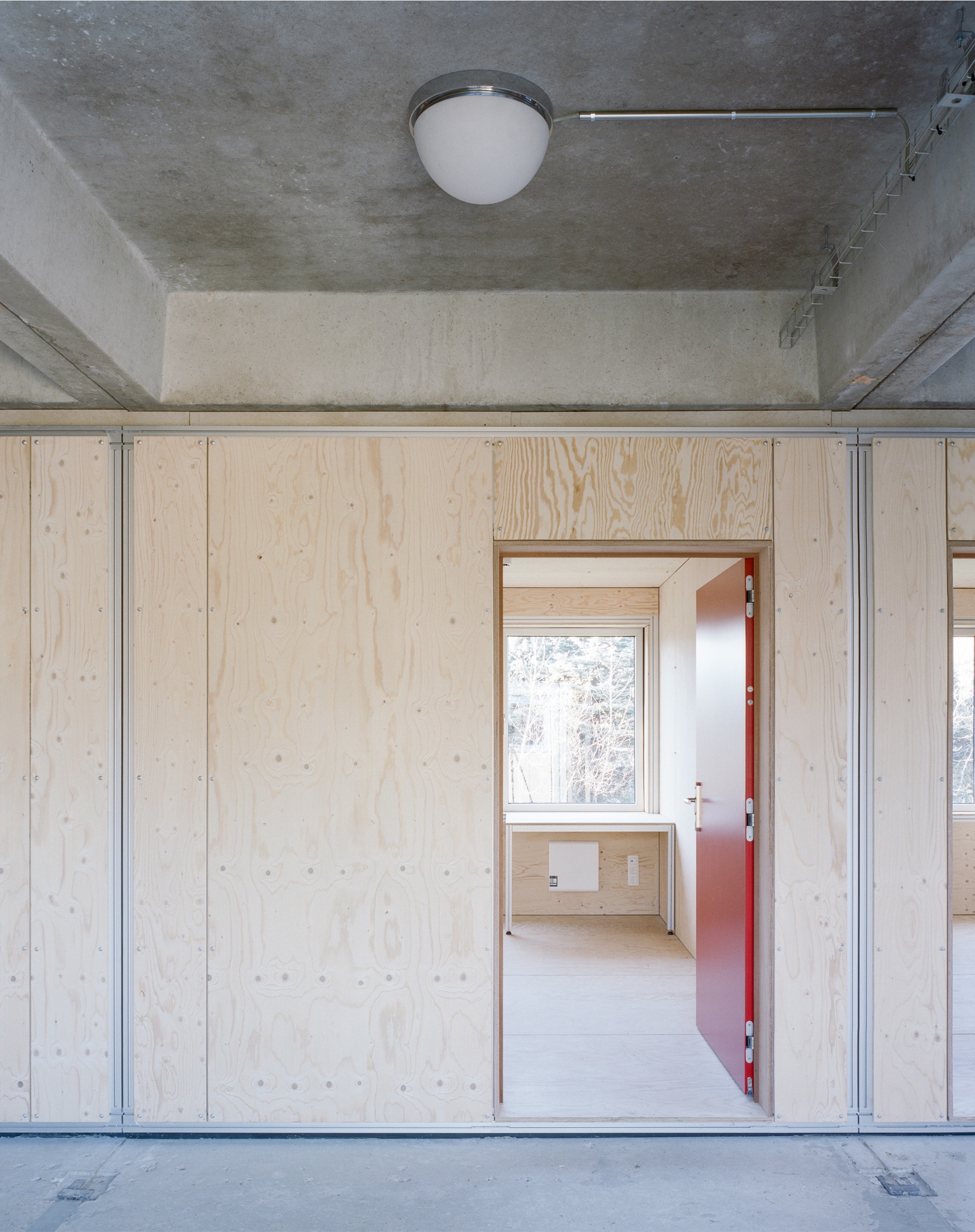 Light plywood wall with salmon-colored door opening to room with exposed concrete ceiling and steel channels