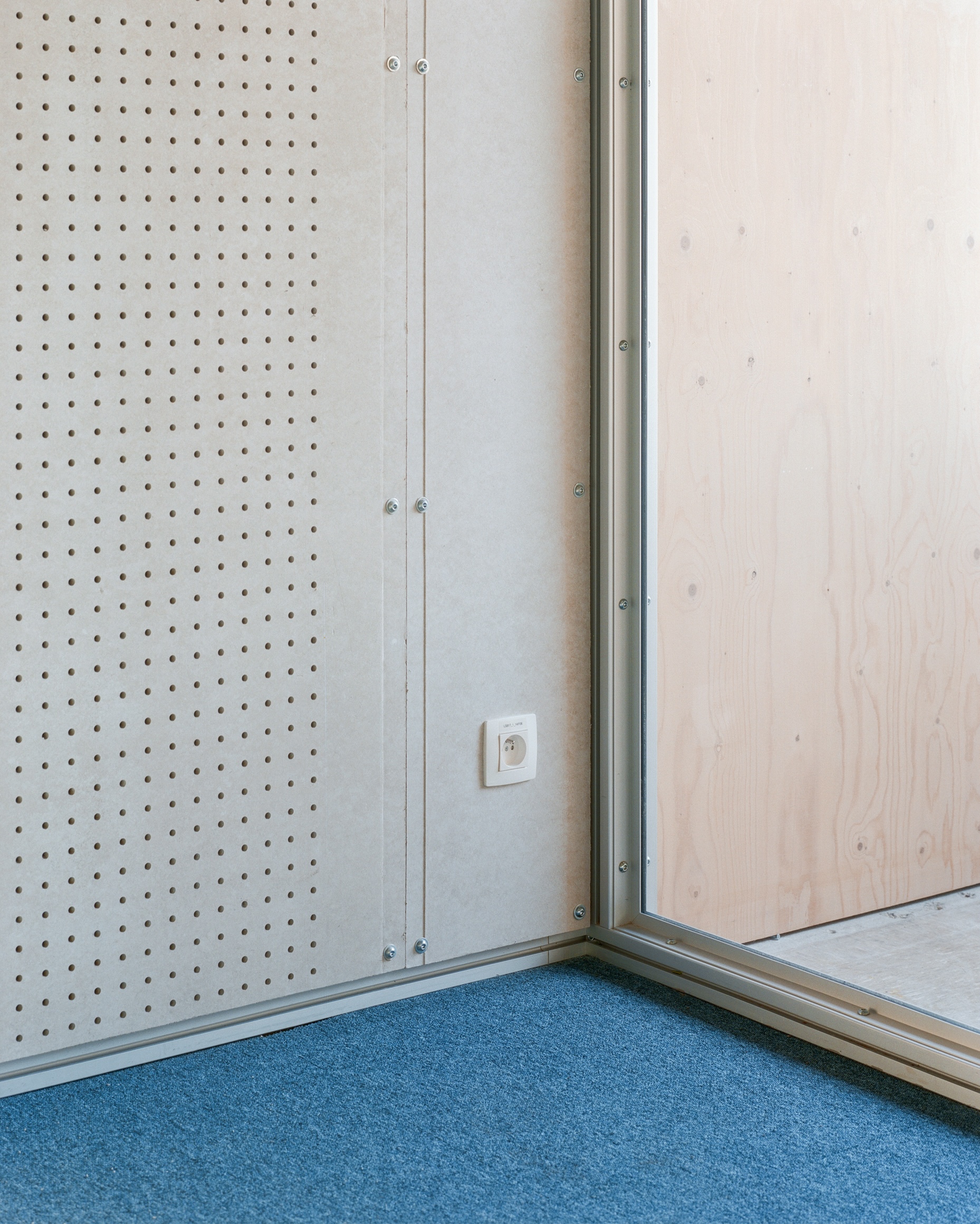Corner junction of perforated plywood panel, blue carpet and glass sliding track