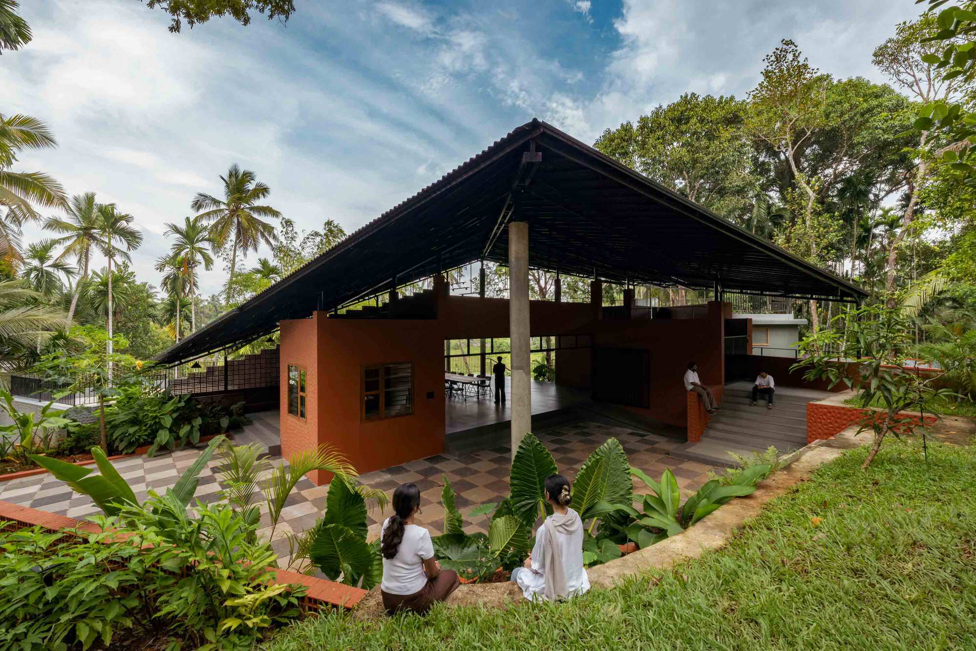 Exterior view of pitched roof pavilion with terracotta walls and planted courtyard with three figures seated