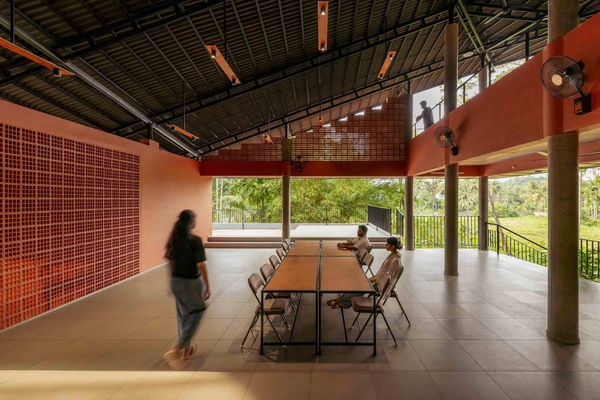 Open pavilion with perforated terracotta screen wall and exposed metal truss ceiling as people gather at a table