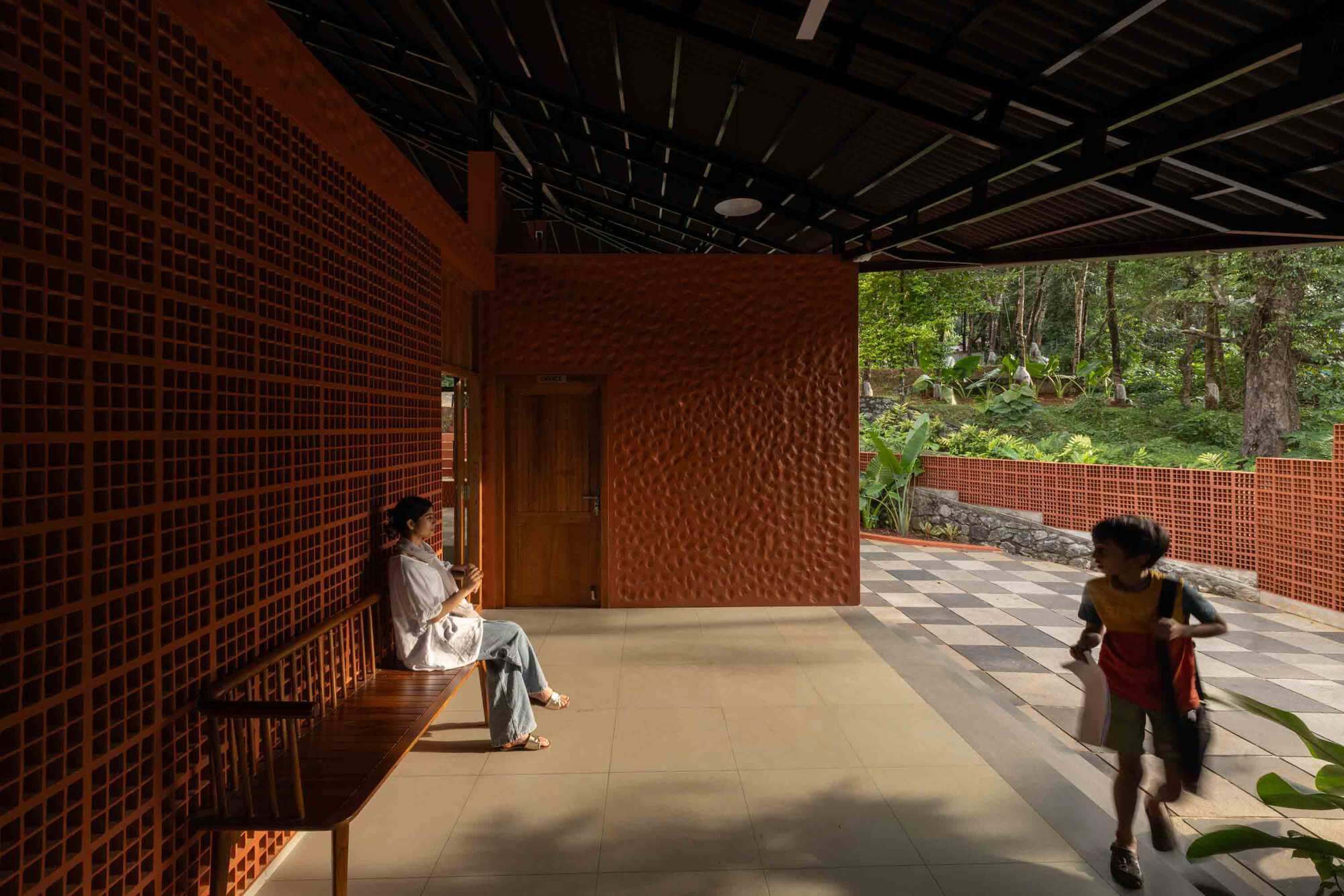 Interior corridor with textured terracotta walls and gridded screening as a woman sits and children play