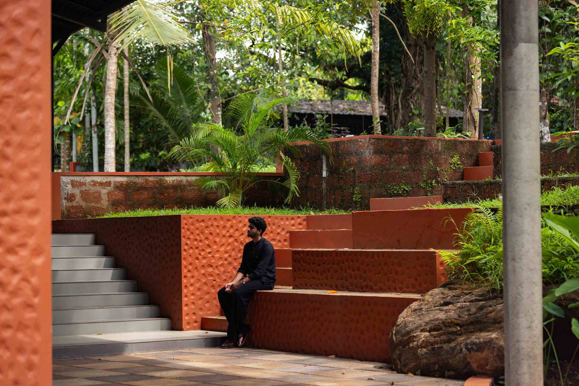 Terraced outdoor steps framed by laterite retaining walls and tropical trees as a person sits beneath the overhang