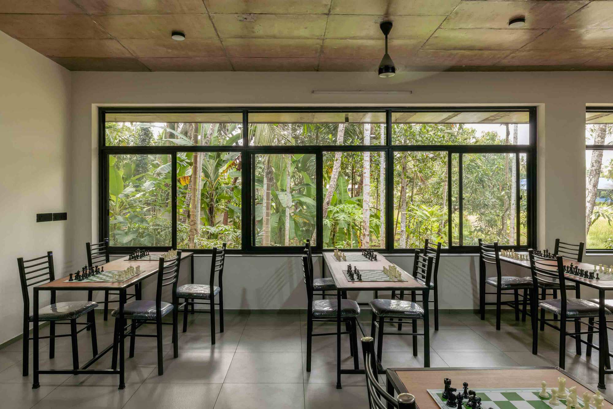 Interior classroom with black-framed windows overlooking dense tropical vegetation and tables arranged for chess