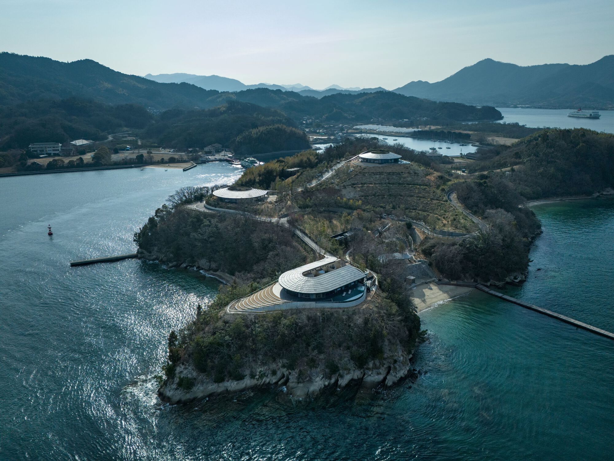 Aerial view of an oval pavilion sited on a forested promontory between two bodies of water