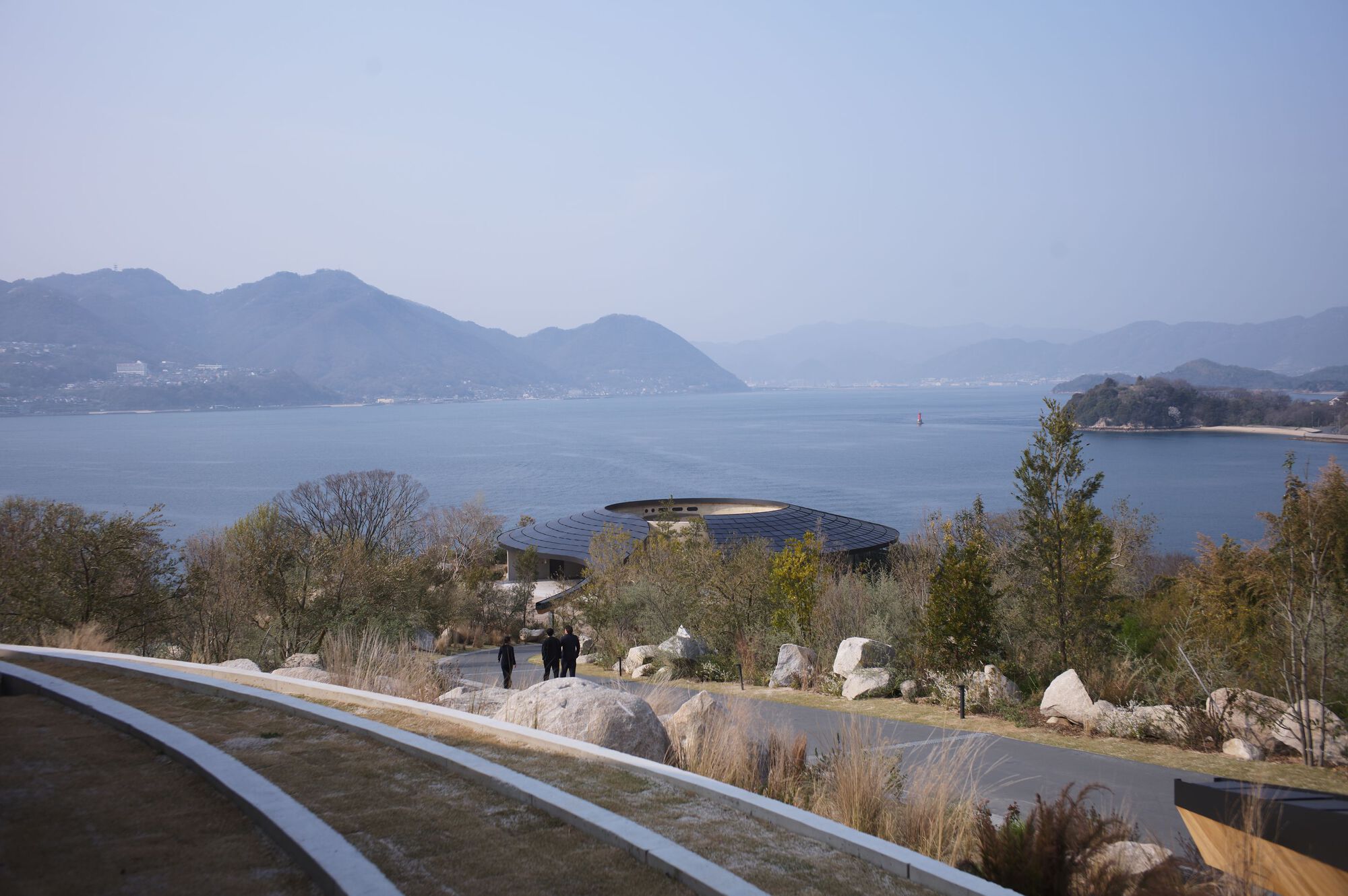 Curved roof pavilion glimpsed across a rocky planted slope with visitors walking amid native grasses