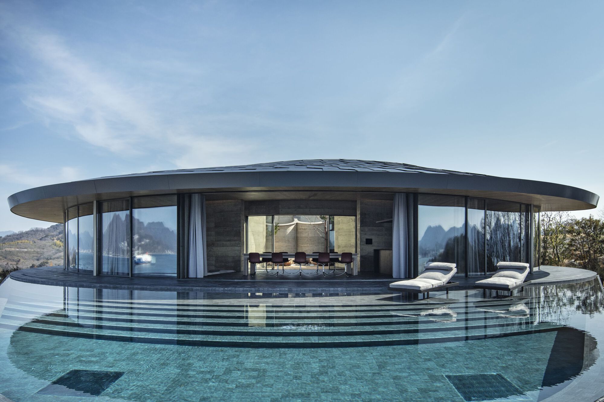 Glass-walled dining pavilion with floating roof canopy and infinity pool reflecting the mountains beyond