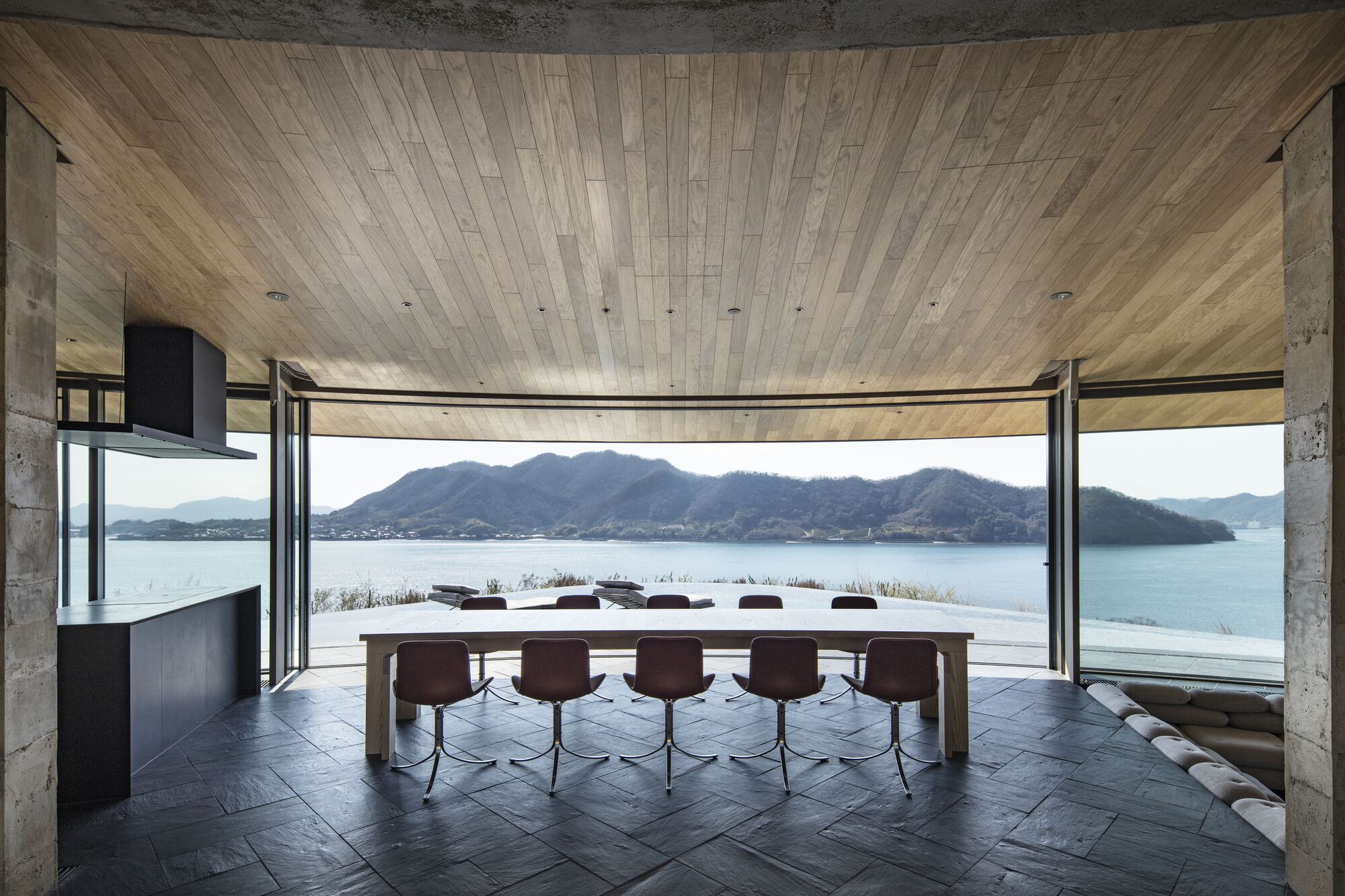 Interior dining space with curved timber ceiling, floor-to-ceiling glazing and long table overlooking the water