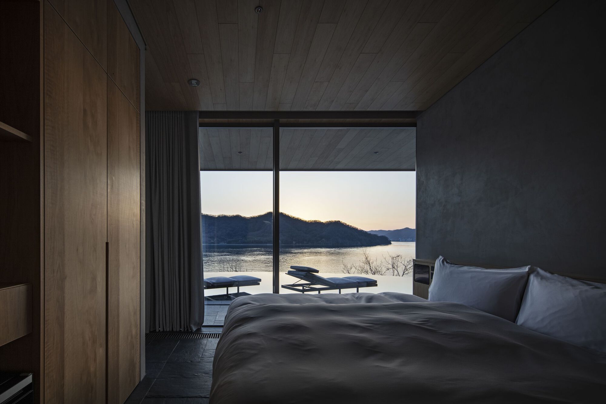 Bedroom with timber ceiling and concrete wall framing a lakeside view at dusk