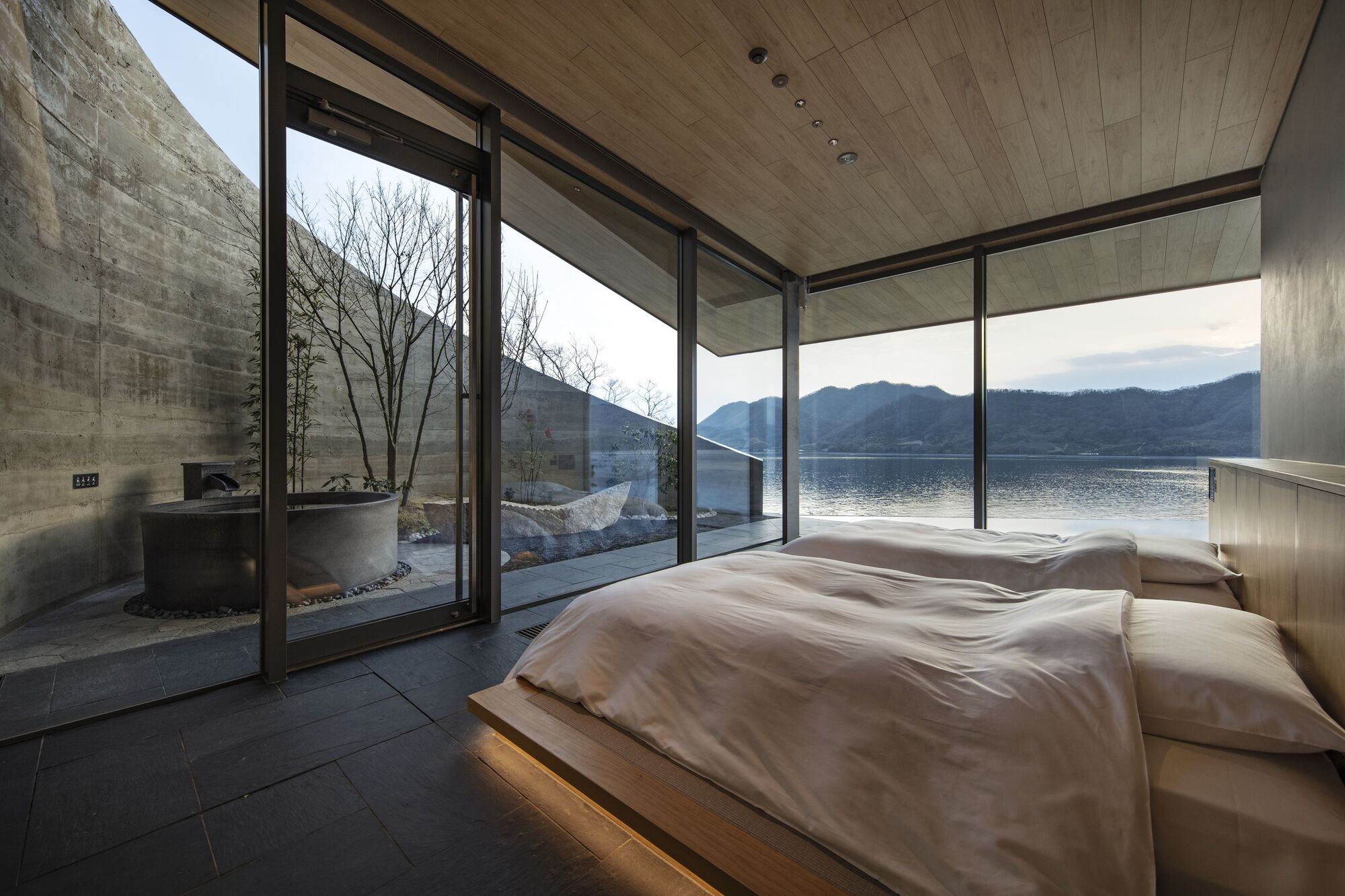 Floor-to-ceiling glazing opening the bedroom to a terrace with bare winter trees and lake view