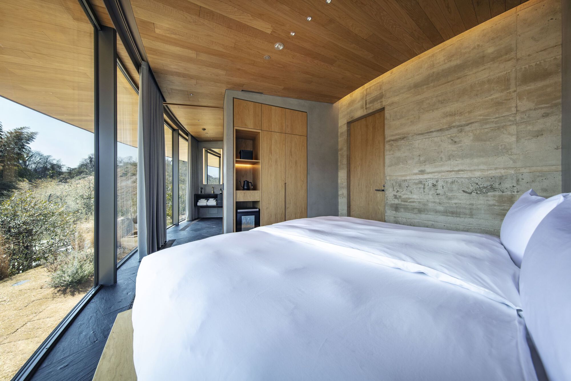 Bedroom with board-formed concrete walls and glazed corner opening to hillside vegetation
