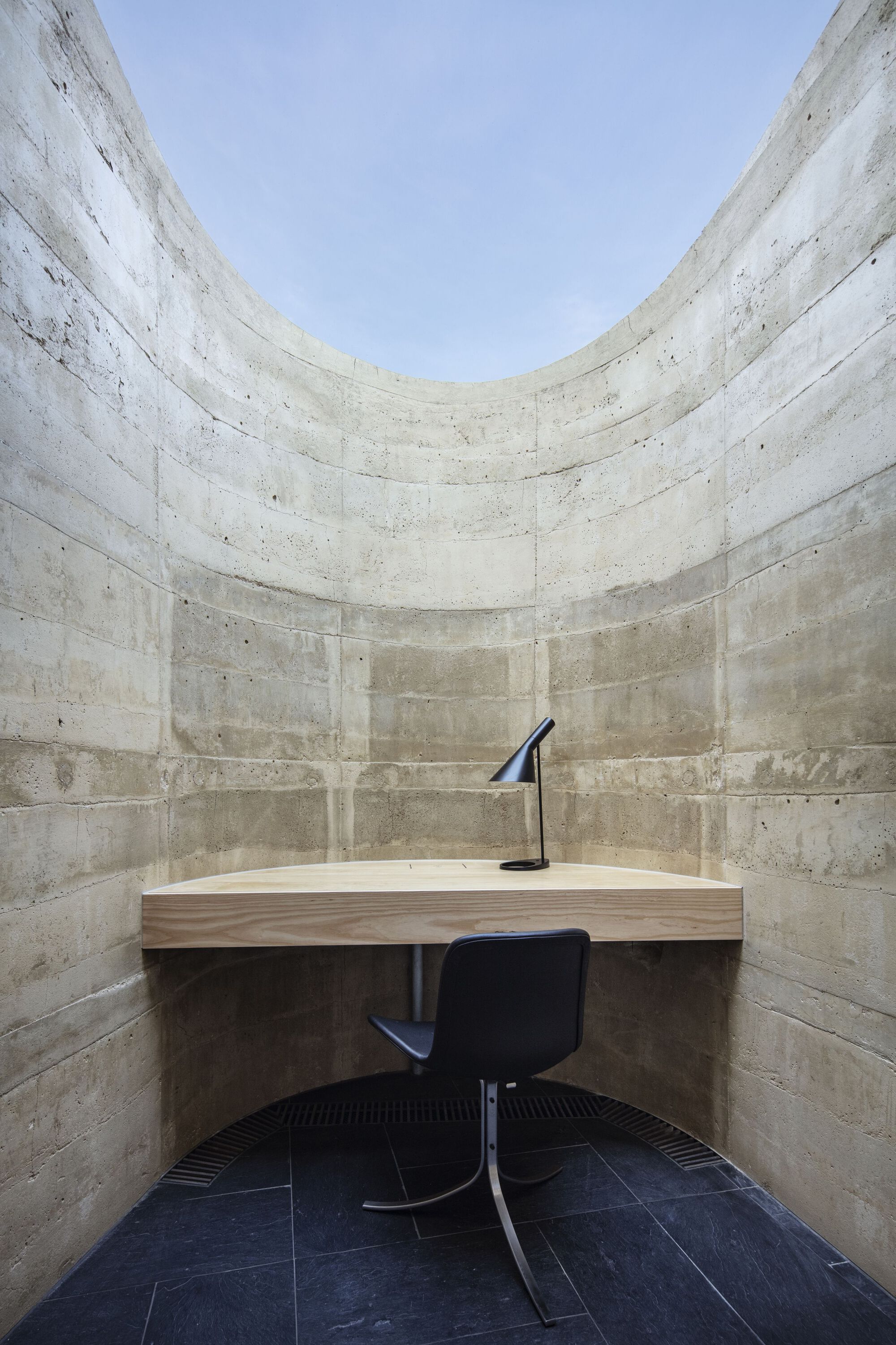 Small study alcove with board-formed concrete walls, timber desk and black swivel chair beneath an open skylight