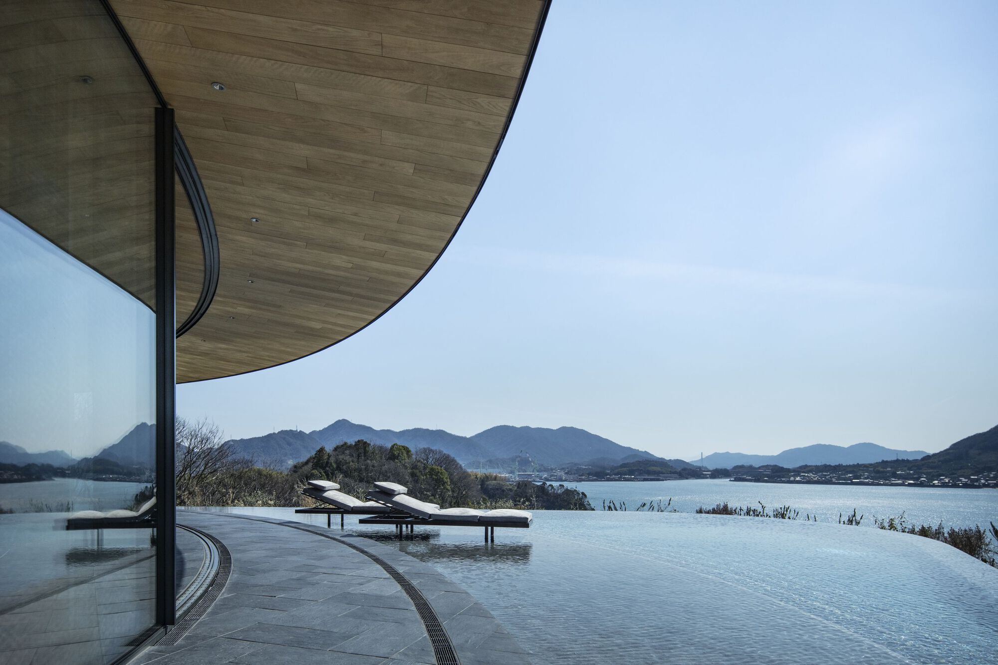Infinity pool terrace with timber ceiling overhang overlooking a lake and distant mountain range at midday