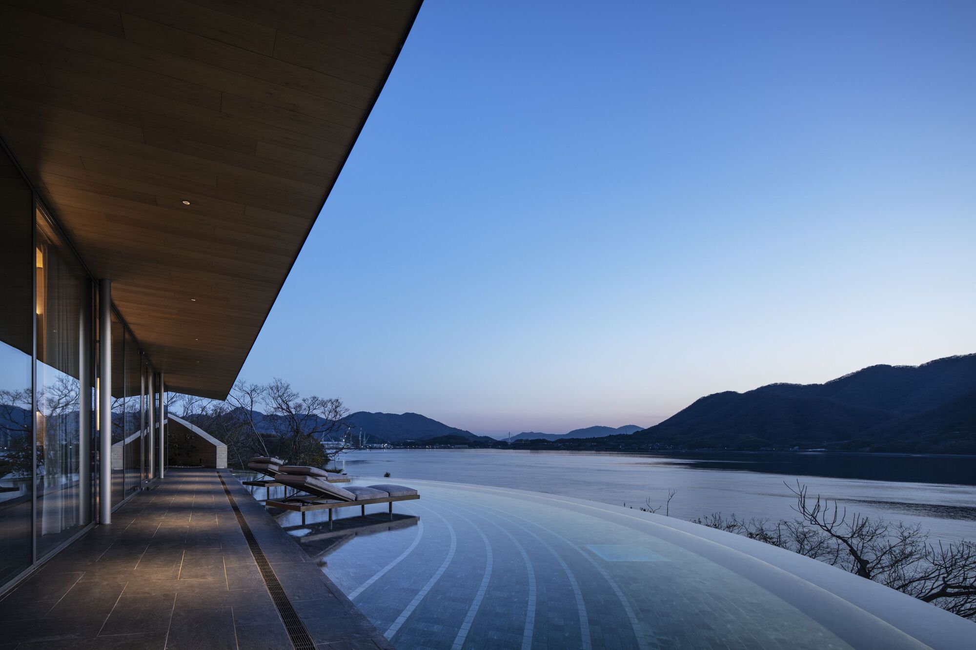 Lakeside infinity pool and glazed colonnade beneath a curved timber soffit at dusk