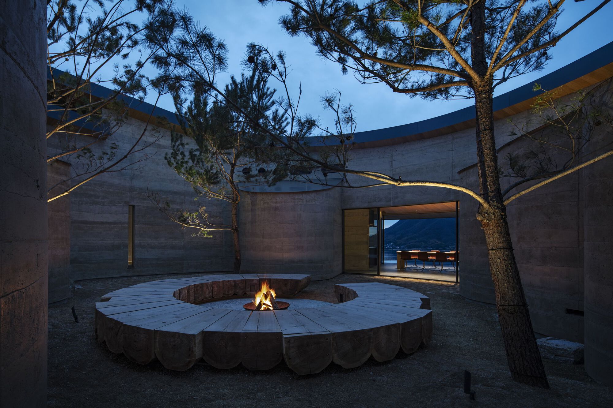 Circular fire pit courtyard with concrete walls, pine trees and glowing interior beyond at twilight