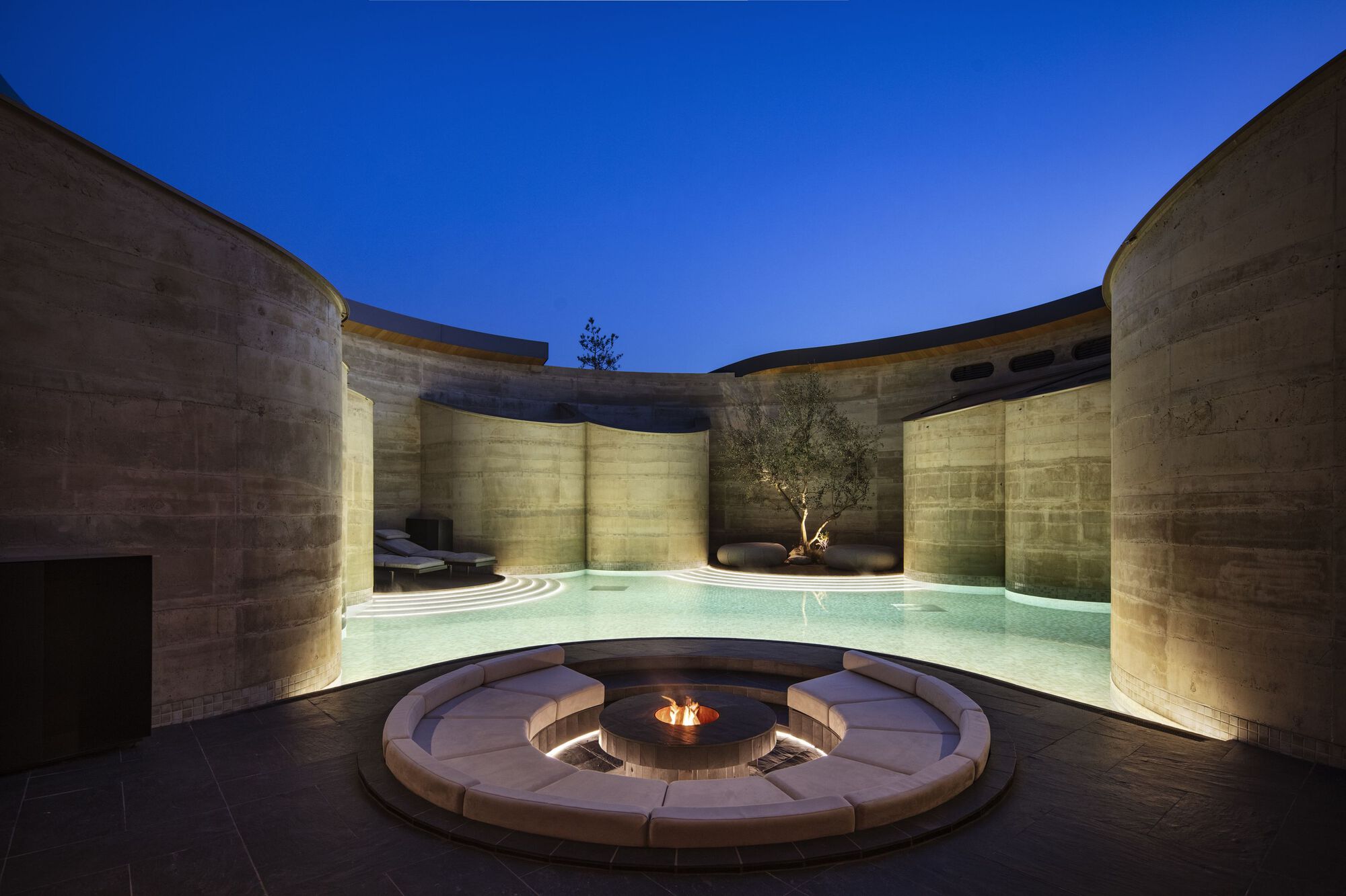 Curved swimming pool and circular fire pit seating area enclosed by uplighting concrete walls at night