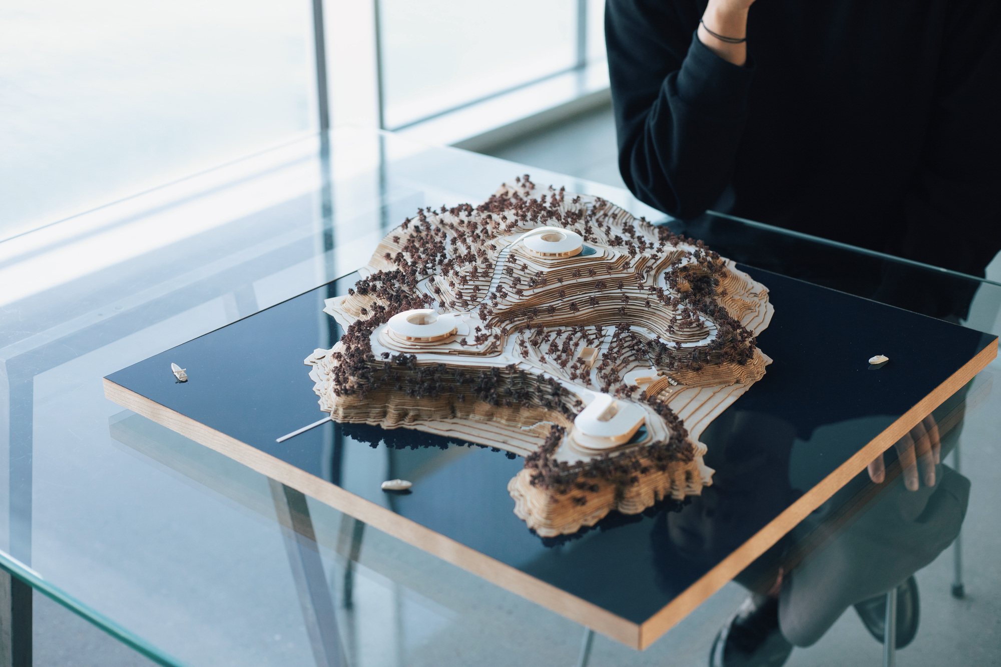 Physical model showing terraced volumes with layered contours and miniature trees on a glass display table