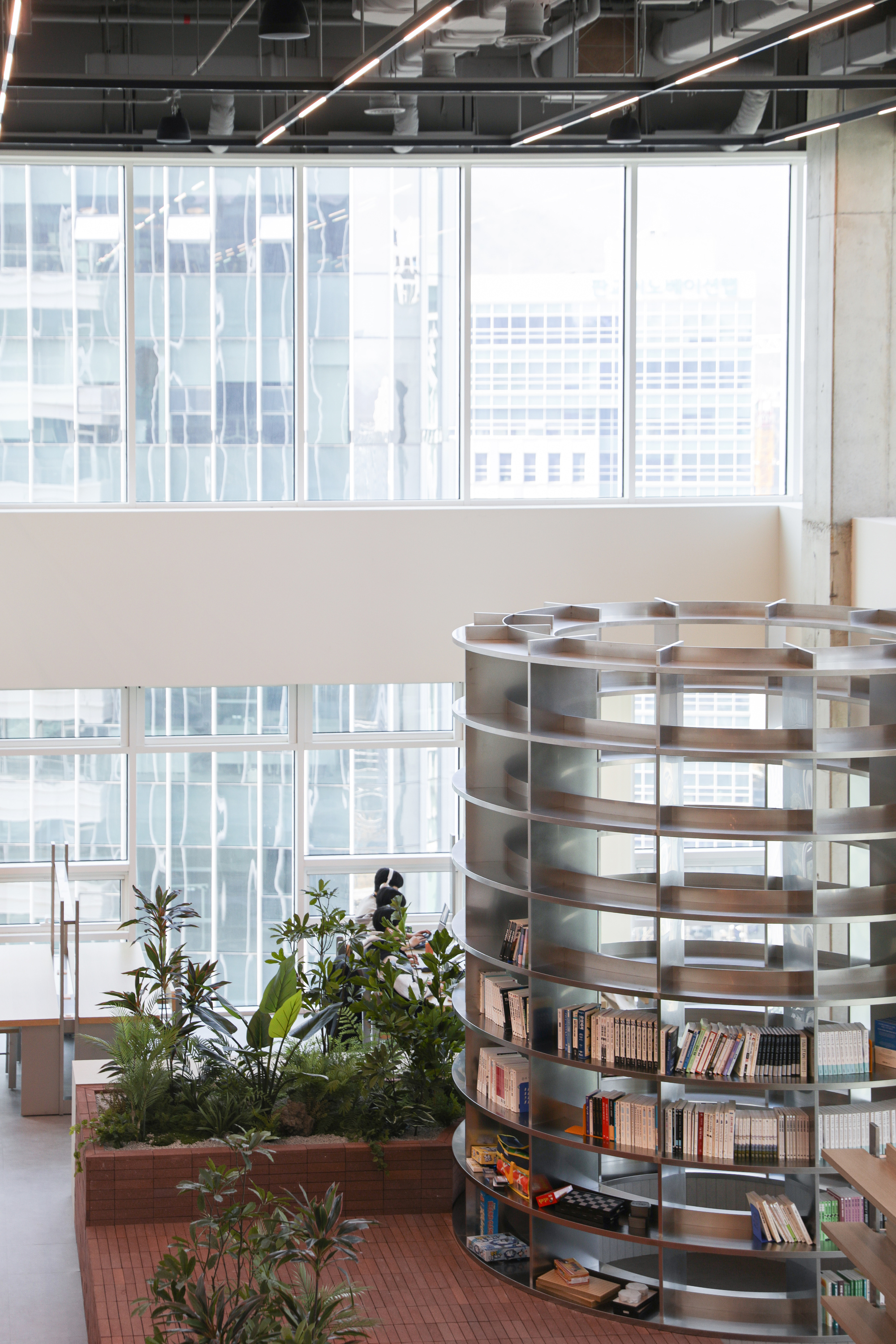Spiral bookshelf beside planters in a double-height space with exposed ceiling services and curtain wall windows