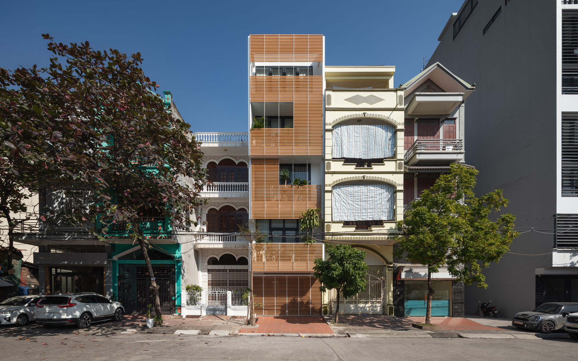 Street view of a narrow tower with horizontal timber louvers flanked by neighboring buildings under a clear blue sky