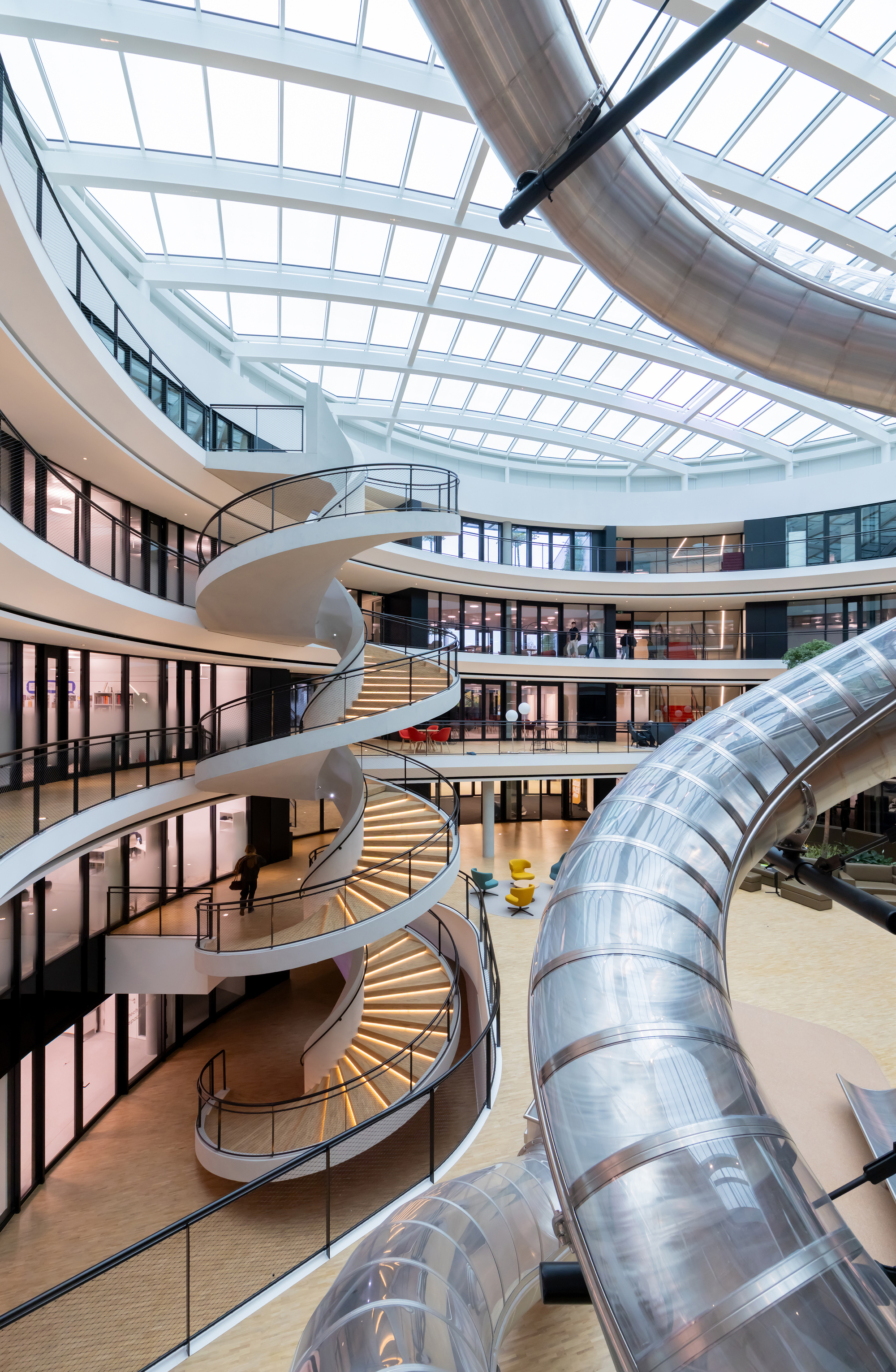 Multi-story atrium with spiraling staircase and large metal ductwork under a glazed skylight
